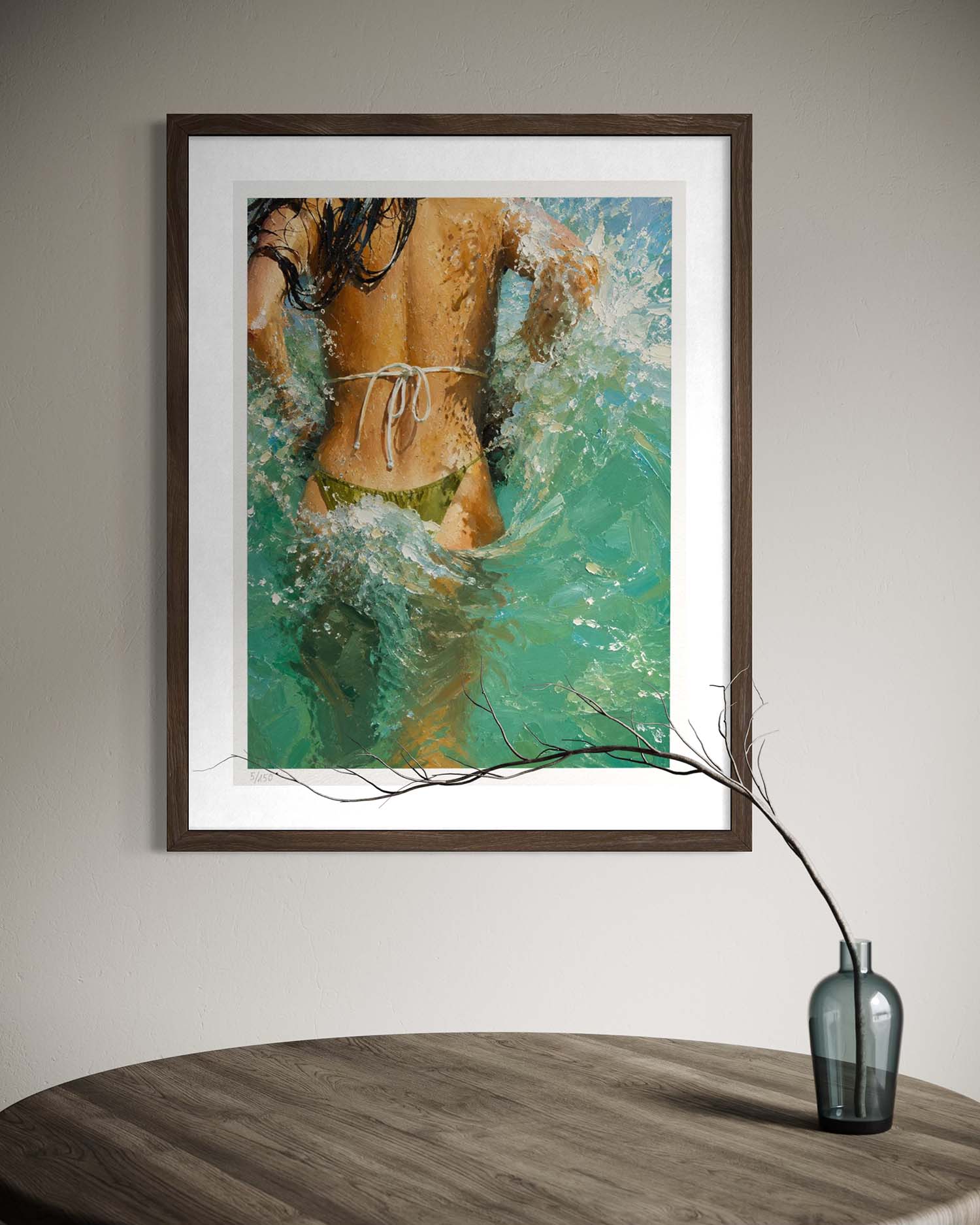 Framed wall art showing a woman from behind in turquoise water, hung on a light gray wall above a round wooden table with a small vase and branch.