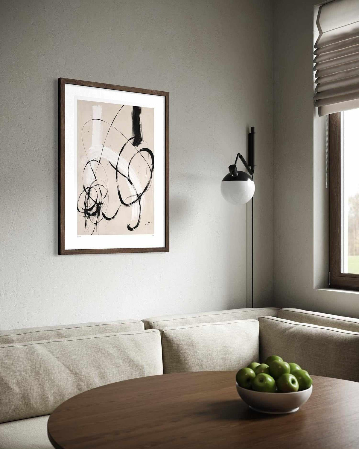 Framed abstract wall art with black curved brushstrokes on a warm beige background, displayed above a light beige sofa in a modern living room setting with a wooden table and bowl of green apples.