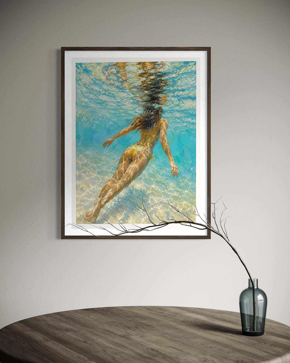 A framed art print depicting an underwater scene of a woman swimming, displayed on a light gray wall above a wooden table with a glass vase.