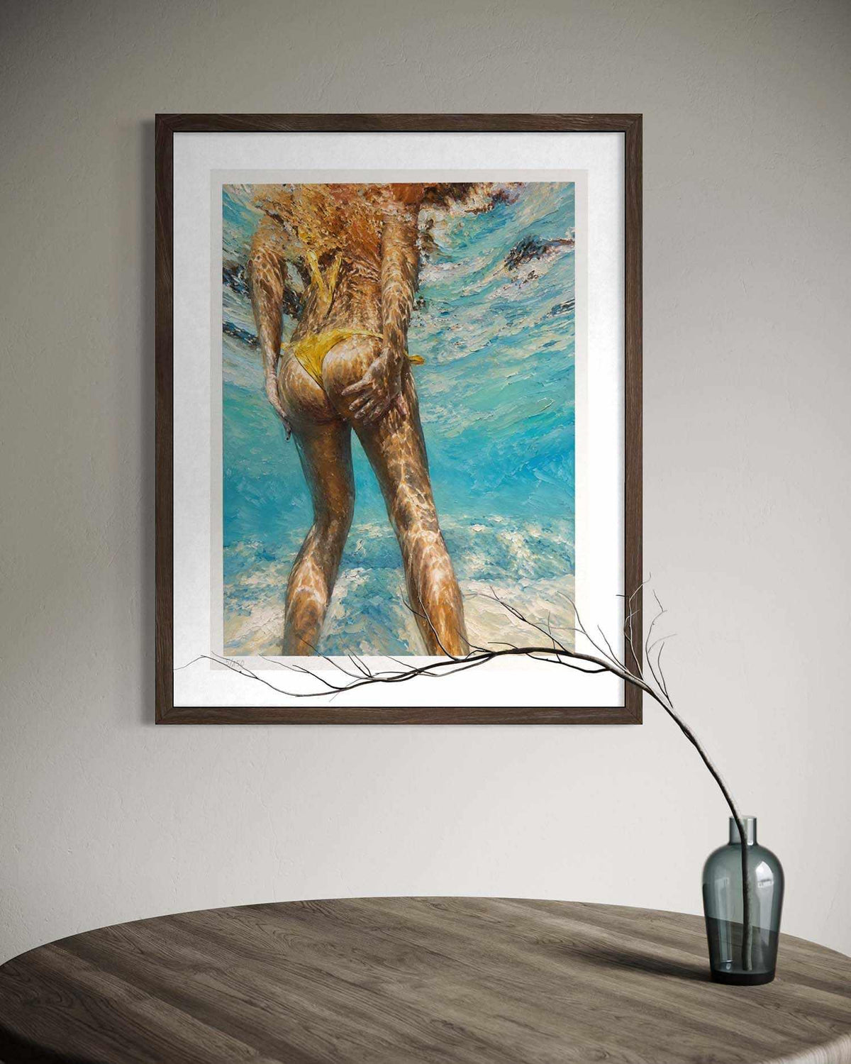 A framed art print depicting a woman's back underwater in a yellow bikini, hanging on a light gray wall above a wooden table with a glass vase.