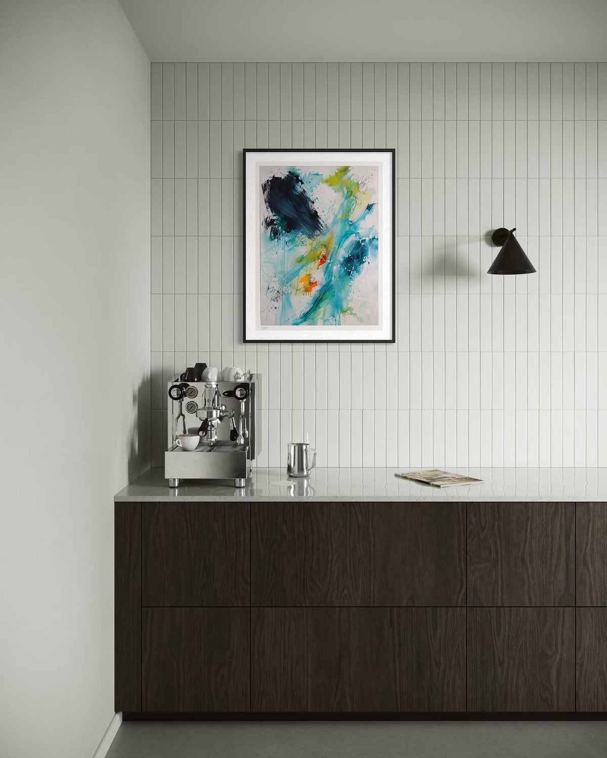 A framed abstract art print with blue, green, yellow, and black brushstrokes hangs on a white tiled wall above a dark wood counter with a coffee machine and a glass.