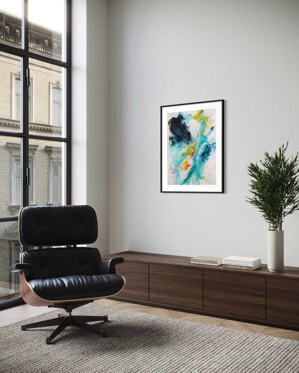 A framed abstract art print with blue, green, yellow, and black brushstrokes hangs on a light gray wall in a modern room setting with a black leather lounge chair, a wooden sideboard, and a potted plant.