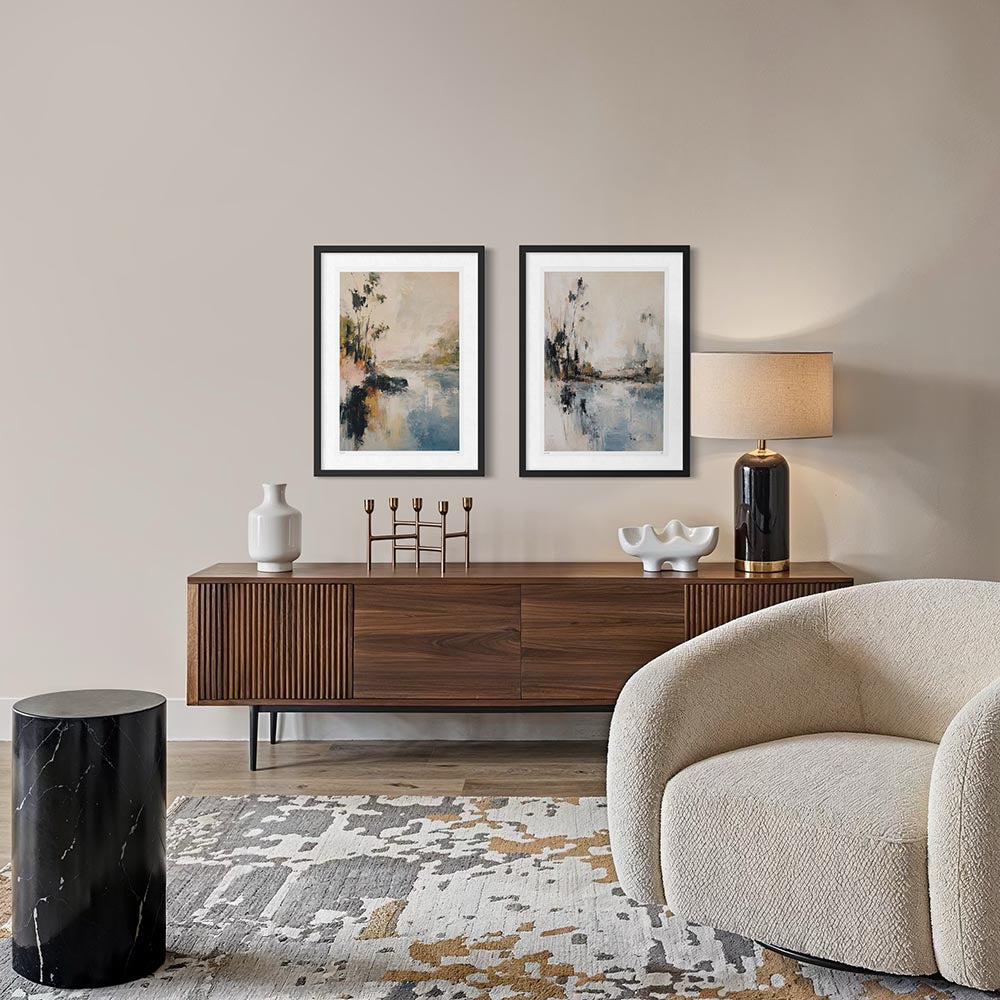 A styled interior scene showing two framed abstract landscape prints hanging above a dark wood sideboard, with a lamp, vase, and decorative objects on the console. A cream upholstered armchair is visible in the foreground, along with a black marble side table and a patterned rug against a warm beige wall.