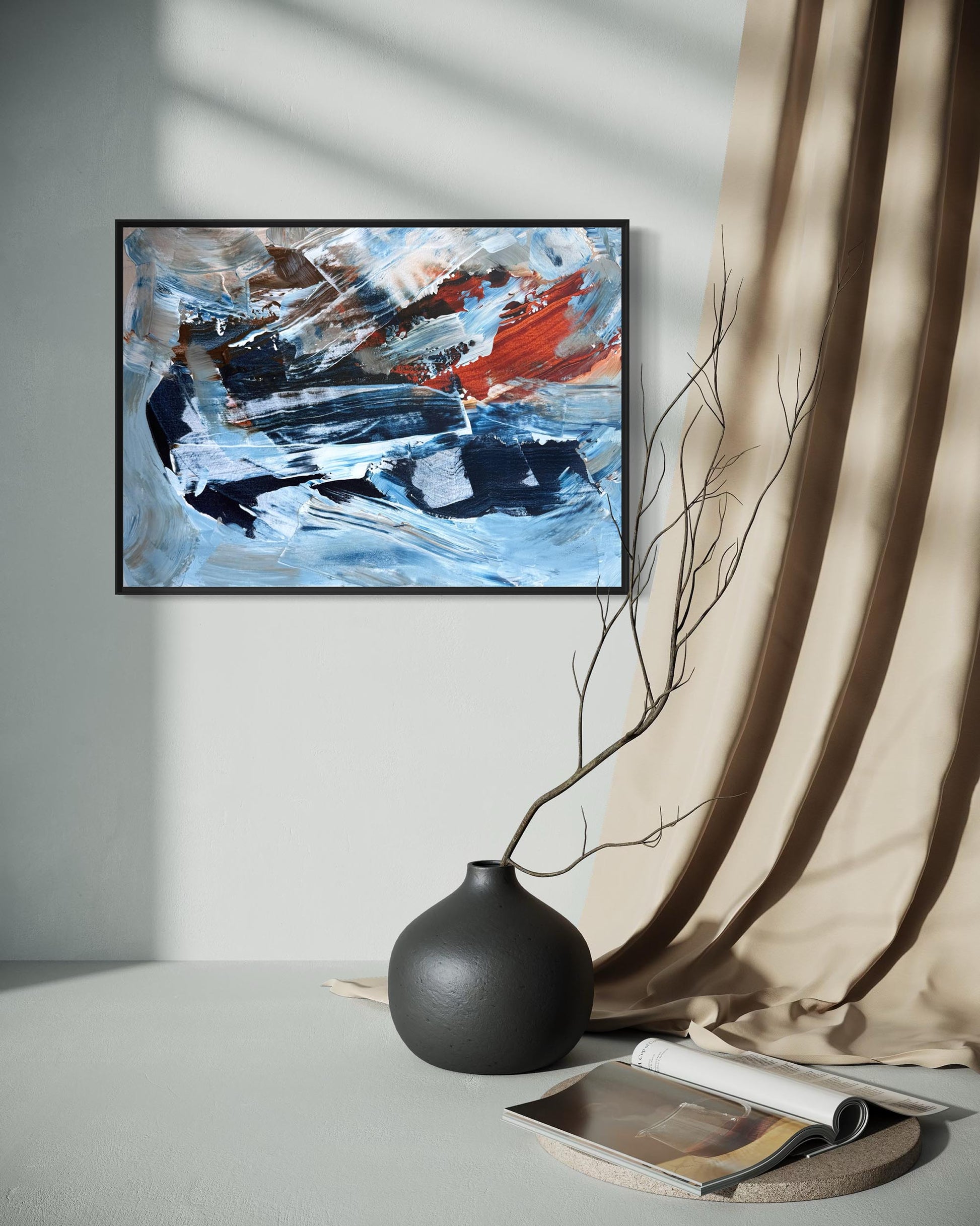 Abstract framed wall art with blue, white, black, and red brushstrokes, displayed in a styled interior next to beige draped fabric, a matte black vase with a bare branch, and an open magazine.