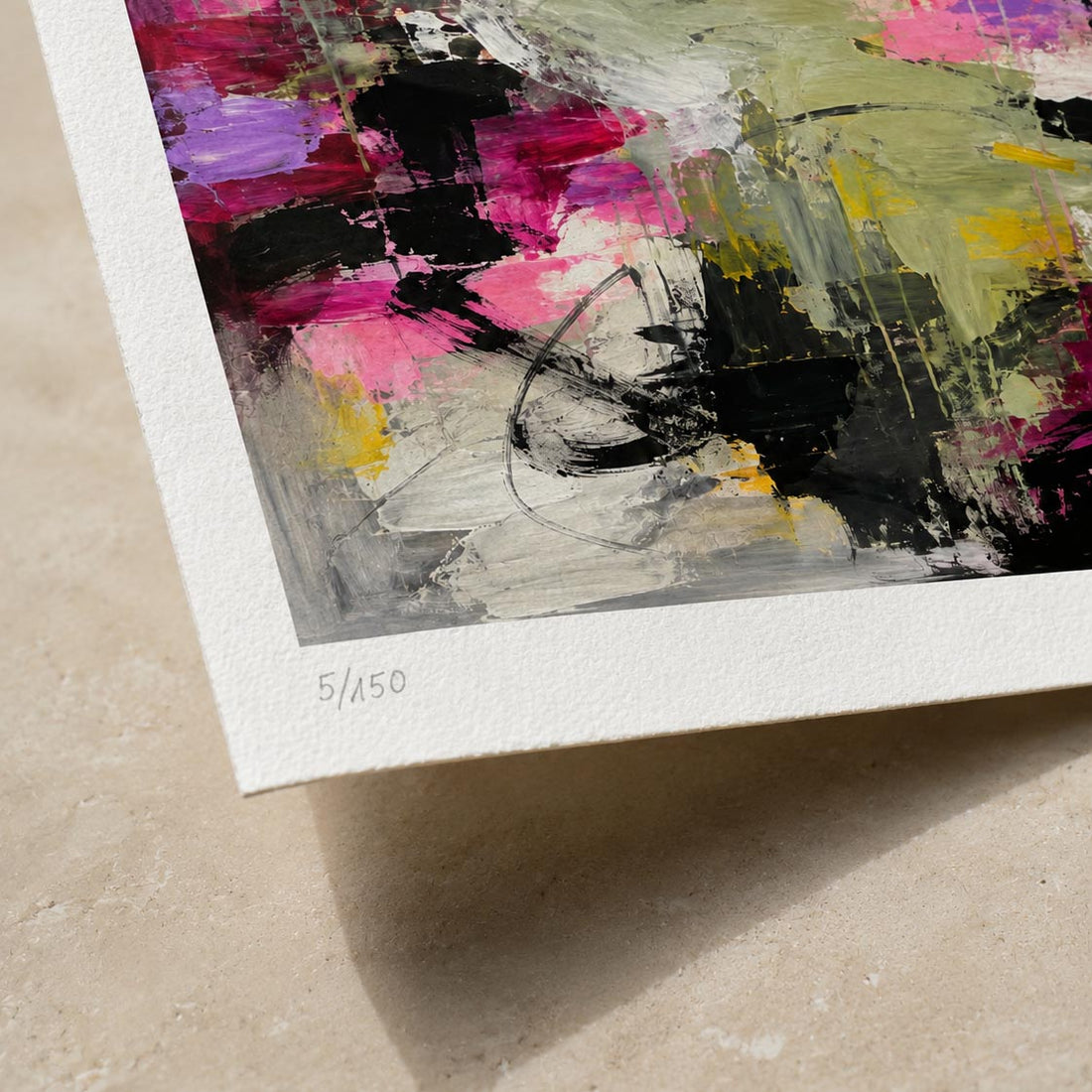 Close-up of an abstract art print on textured paper, showing bold brushstrokes in pink, black, green, yellow, and purple with a white border and the printed edition mark 5/150 on the bottom left, photographed on a beige stone surface.
