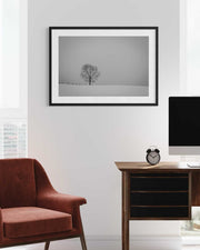 Framed black and white tree photograph on a wall above a brown armchair and wooden desk.