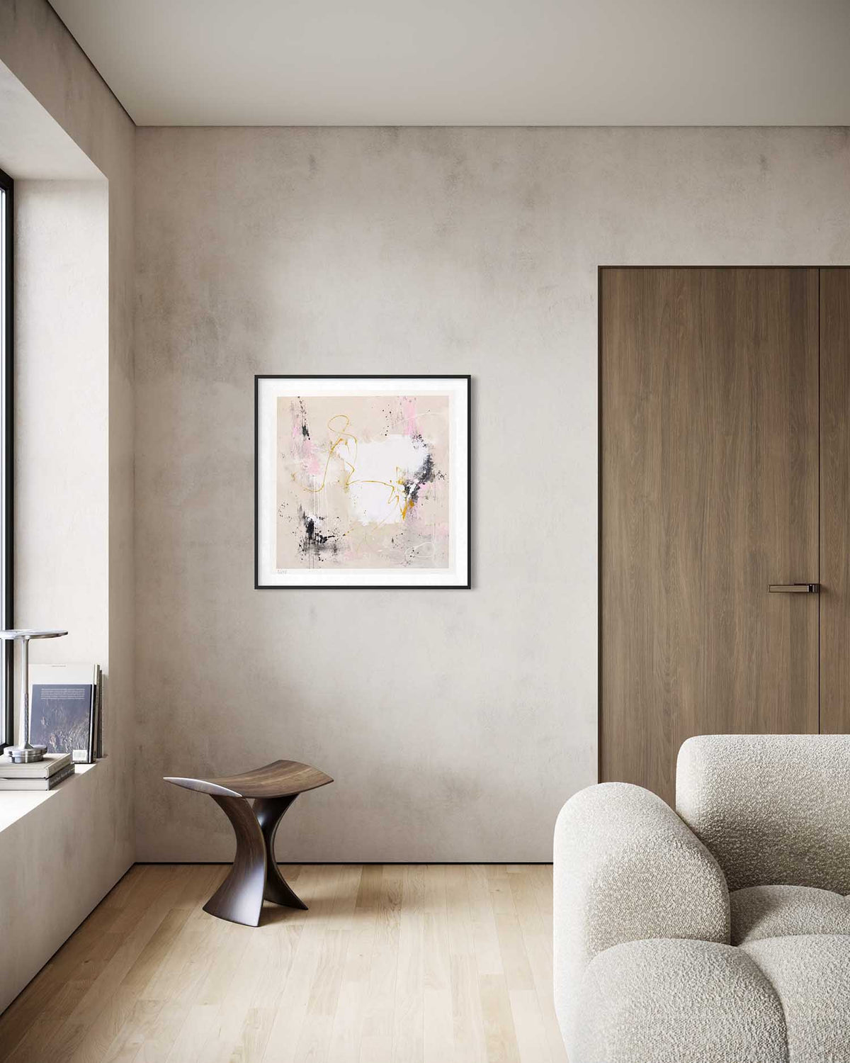 A framed abstract art print with soft pink, white, and gold brushstrokes hangs on a textured beige wall in a modern living room setting with a wooden door, a small sculptural side table, and a textured armchair.