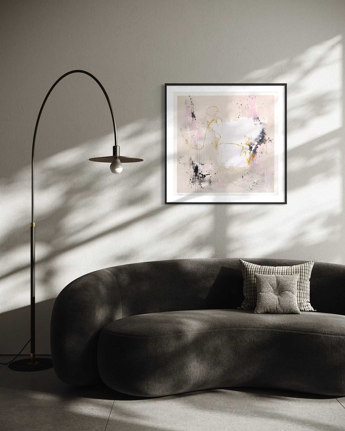 A framed abstract art print with soft pink, white, and gold brushstrokes hangs on a gray wall above a dark gray curved sofa in a modern living room setting with a black floor lamp and dramatic window shadows.