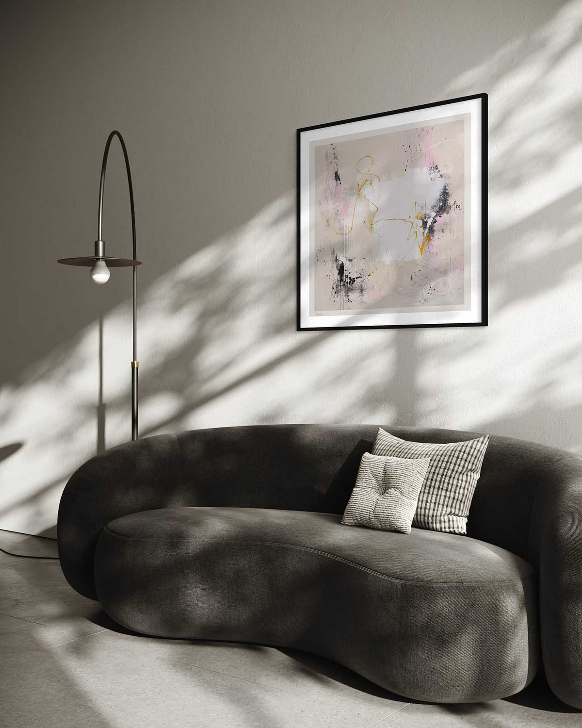 A modern curved gray sofa in a minimalist living room setting, styled with two throw pillows and a framed abstract art print on the wall. A tall, slender black floor lamp stands to the left, casting soft shadows across the sofa and wall.