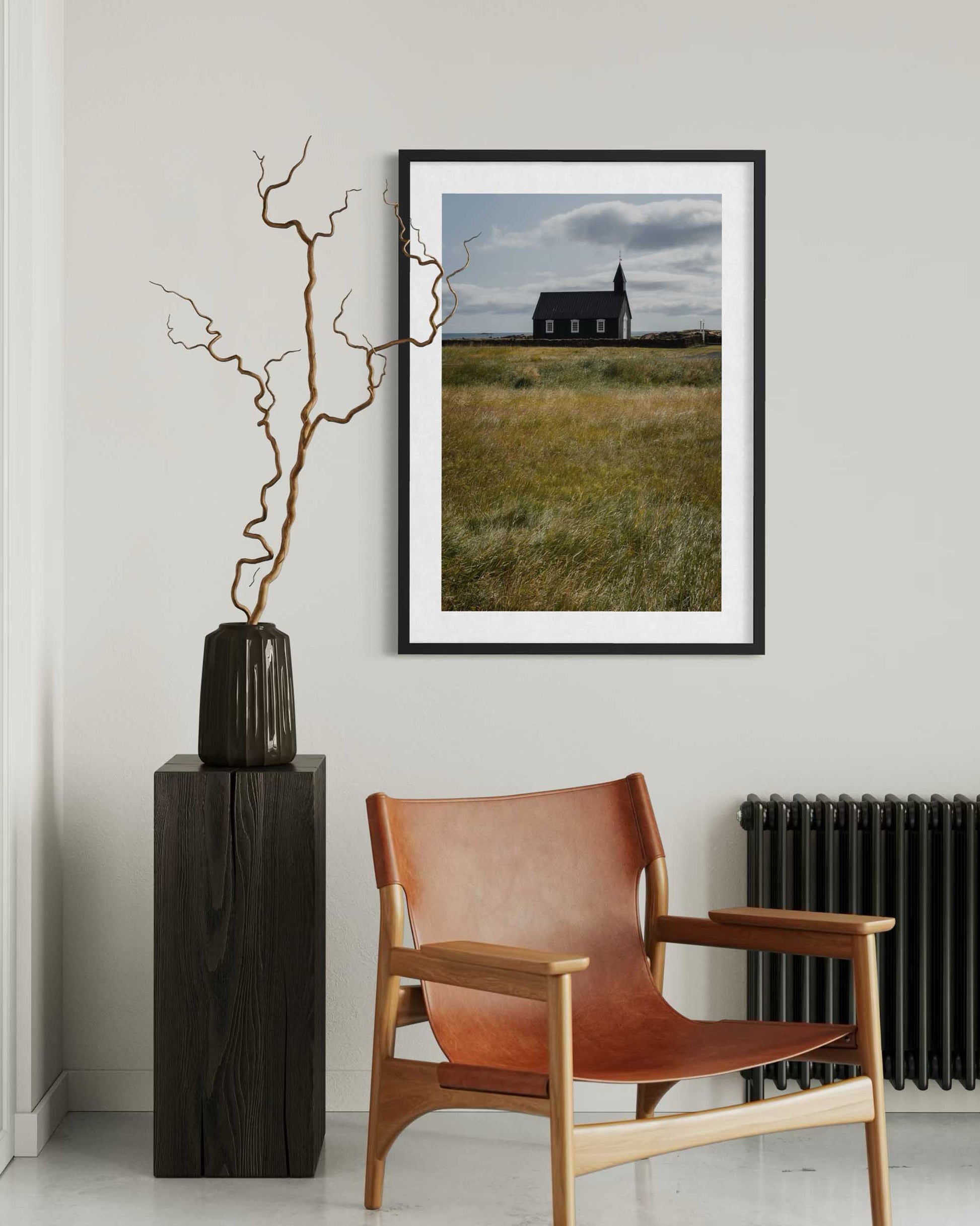 Modern interior with a brown leather chair, vase with branches, and framed landscape art on a white wall.
