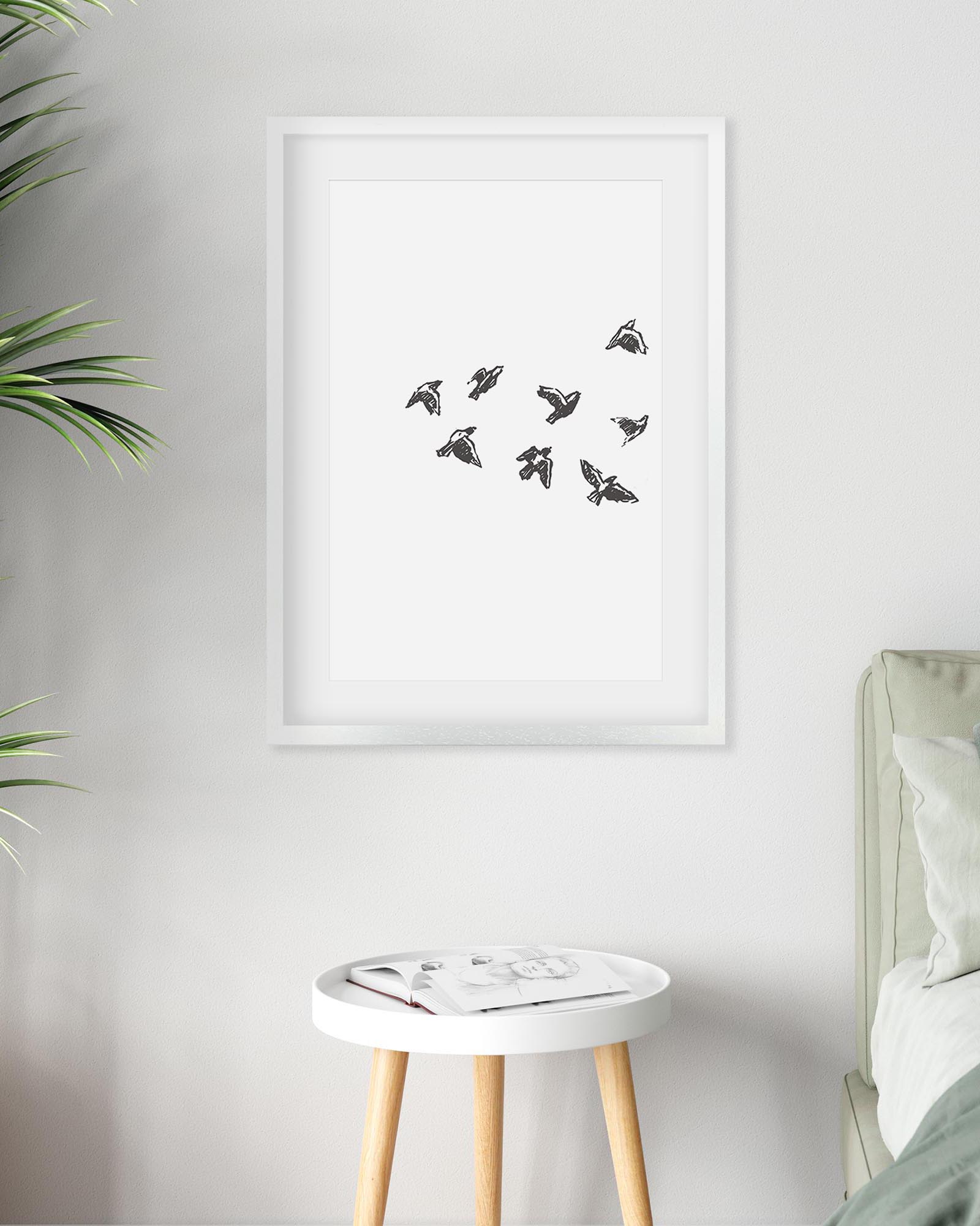 Framed artwork of birds on a white wall above a small table with a plant and bed.