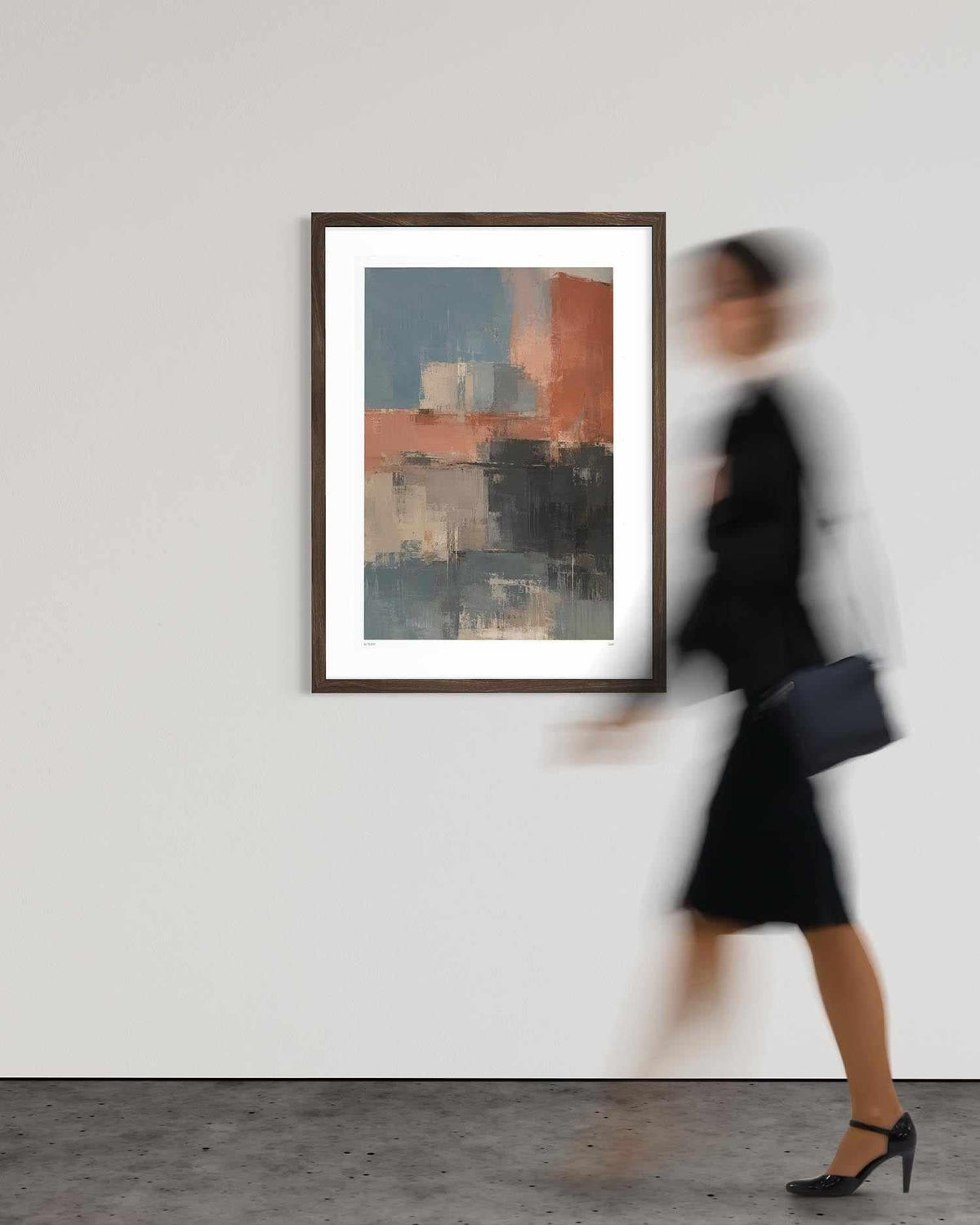 A framed abstract art print with blue, gray, and terracotta brushstrokes hangs on a white gallery wall. A woman in a black dress and heels walks past the frame on the right, creating a motion blur effect.