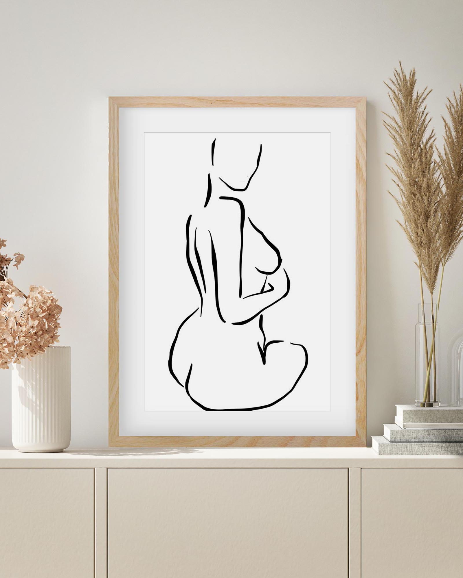 Framed black line art of a nude figure on a white wall with decorative elements.