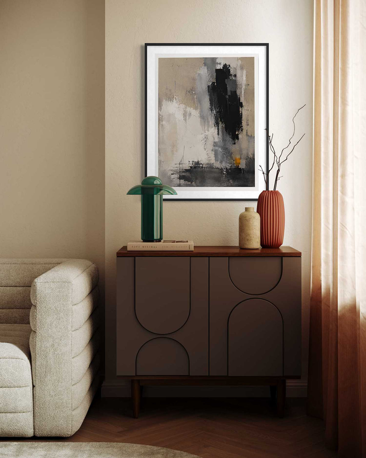 A framed abstract art print with black, gray, and beige brushstrokes hangs on a beige wall above a dark brown wooden credenza in a styled living room setting.