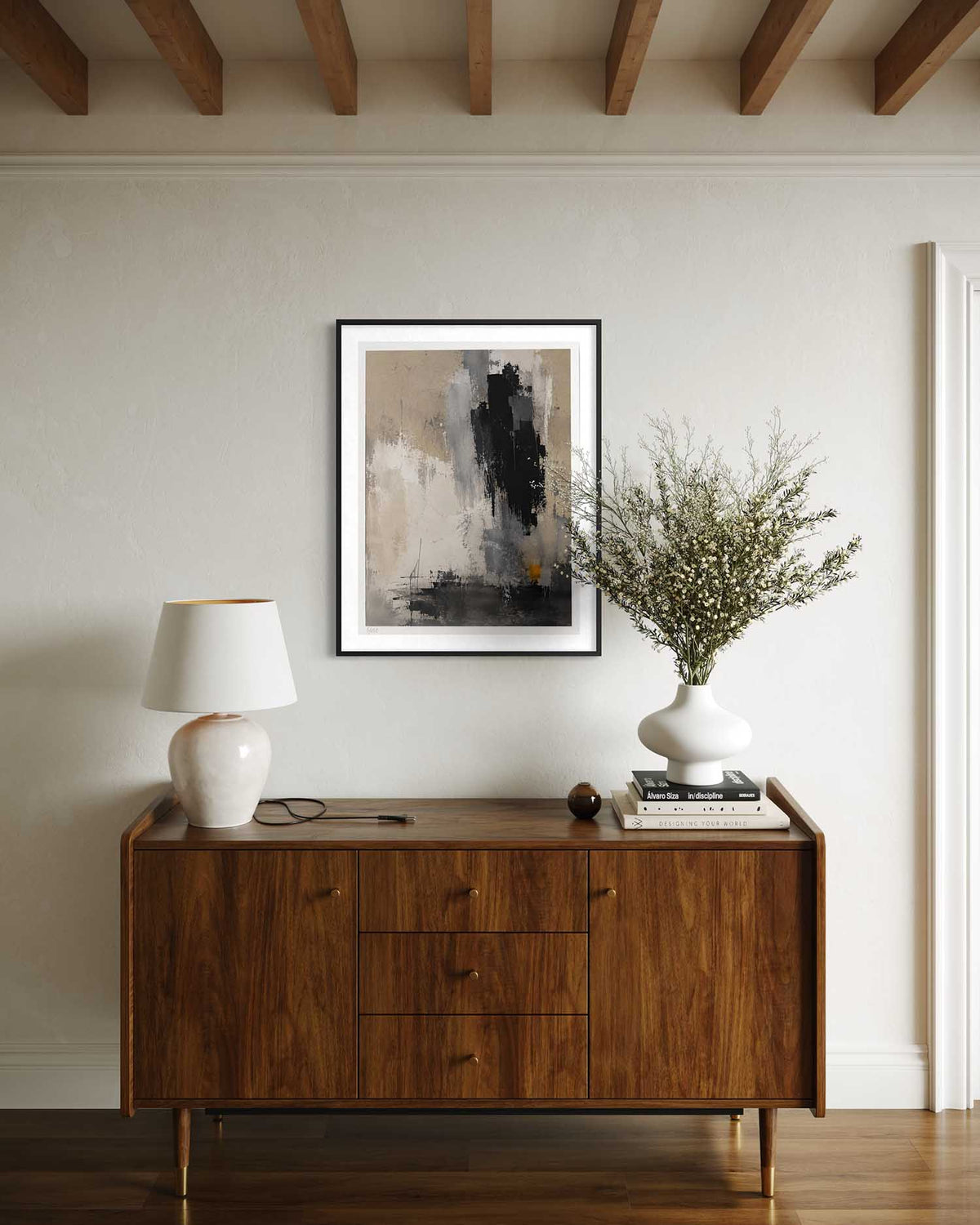 A framed abstract art print with black, gray, and beige tones hangs on a light beige wall above a mid-century modern wooden sideboard. The room is styled with a white ceramic table lamp, a white vase with dried branches, and books.