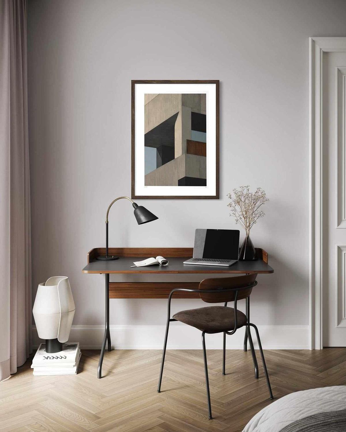 A framed abstract geometric art print hangs on a light gray wall above a wooden desk with a laptop, lamp, and chair in a modern styled room.