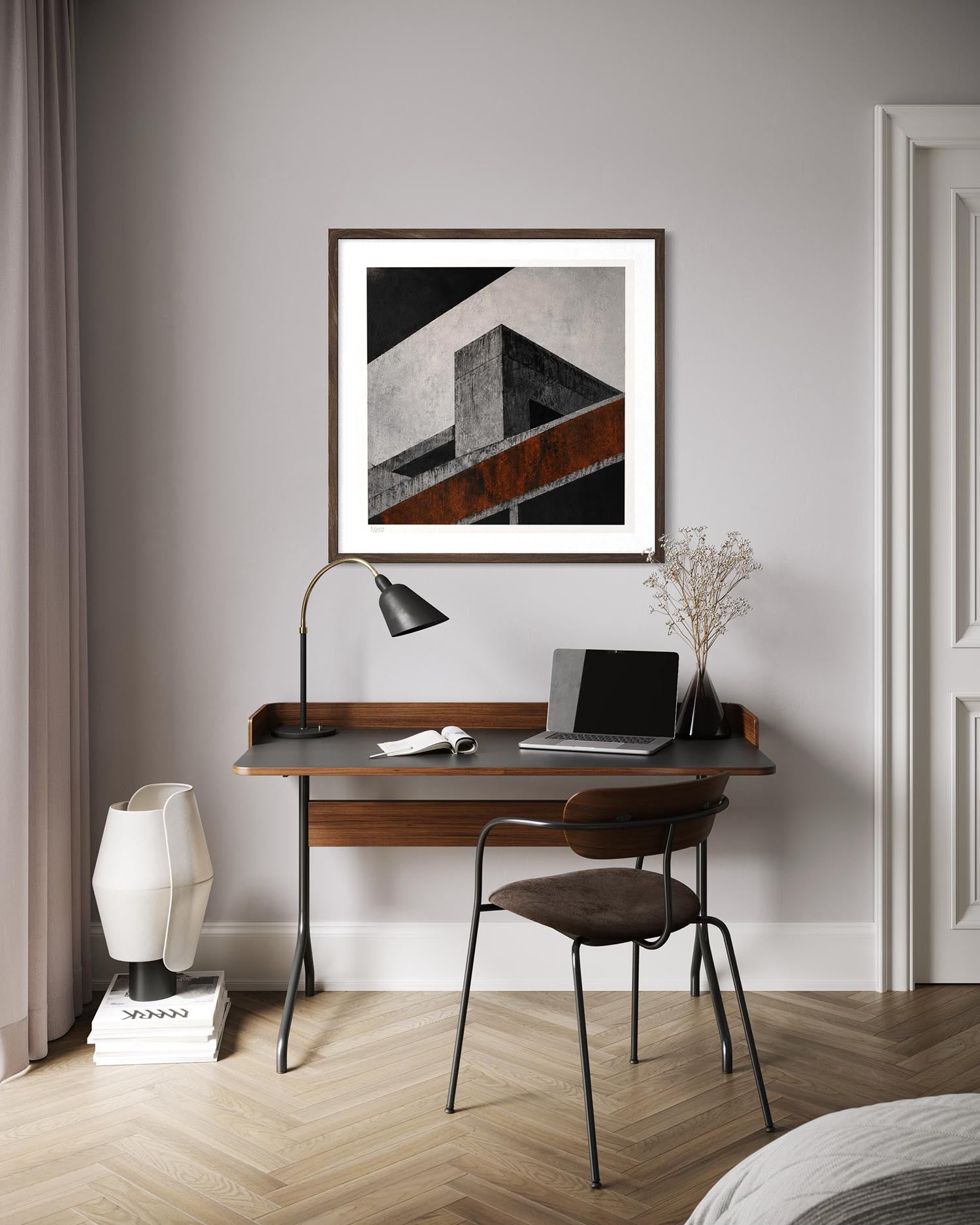 A framed abstract geometric art print hangs on a light gray wall above a wooden desk with a laptop, lamp, and chair in a modern home office setting.