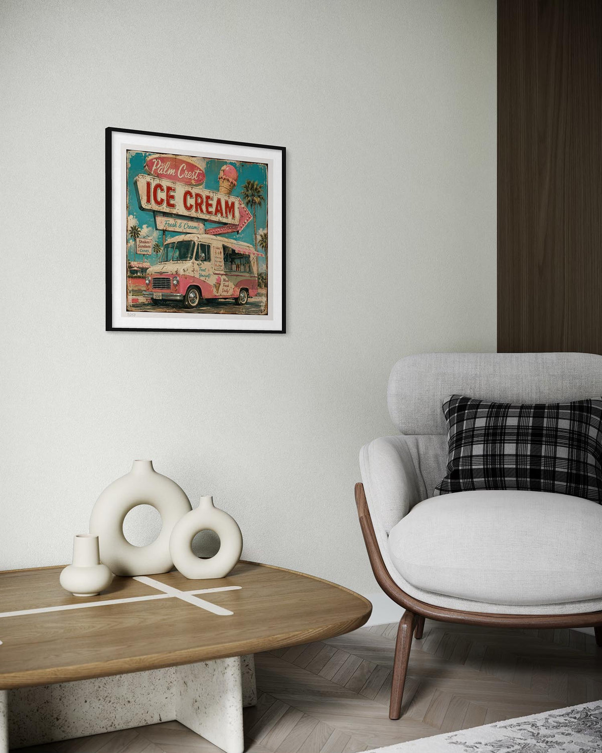 A framed vintage-style ice cream truck poster hangs on a light gray wall in a modern living room setting, next to a white armchair with a black-and-white plaid pillow and a wooden coffee table with white decorative vases.