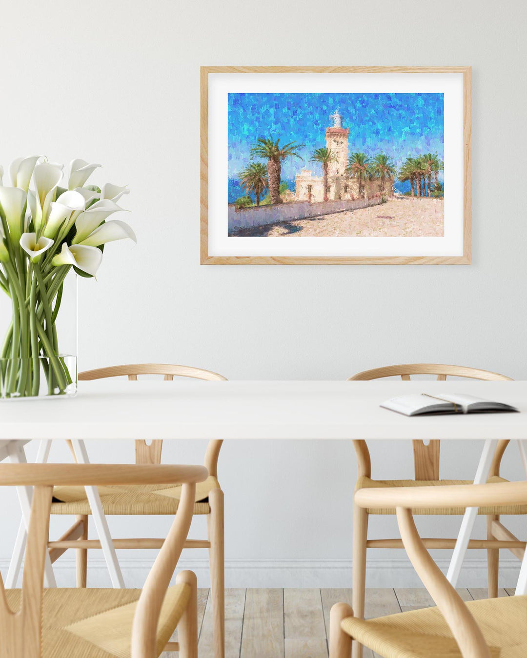 Framed artwork of a coastal scene with a lighthouse on a dining table with flowers and books.