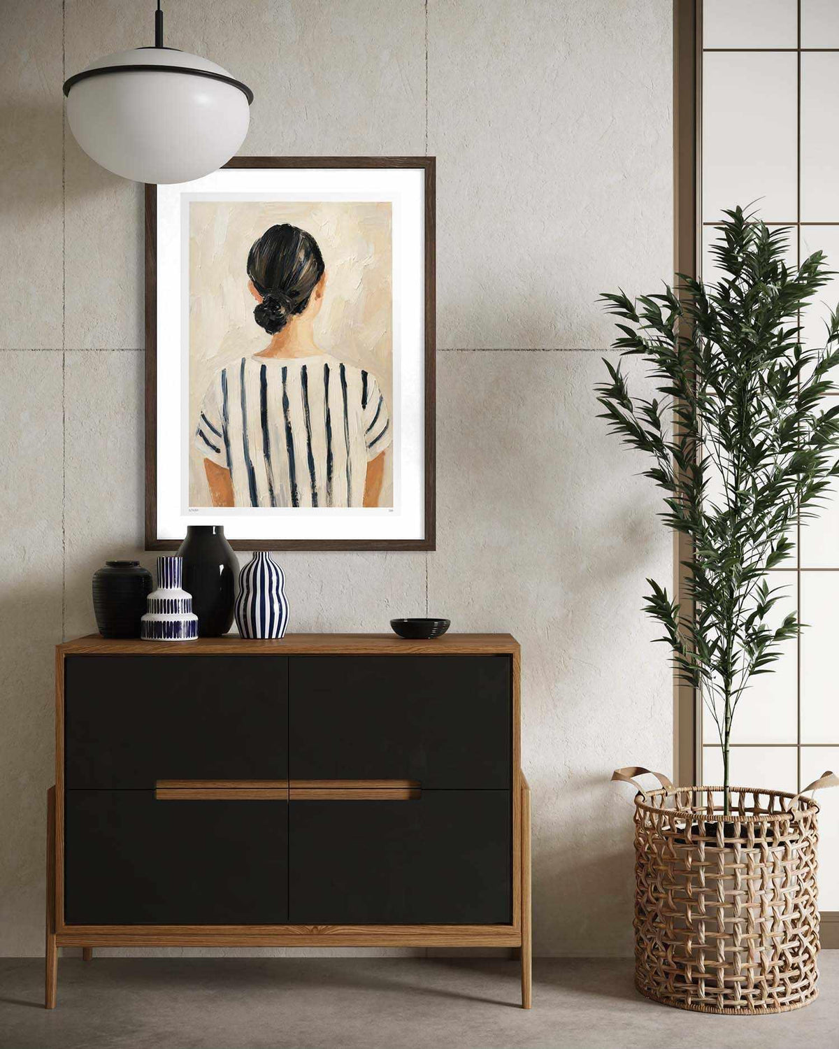 A framed art print depicting a woman from behind wearing a black and white striped top, styled on a beige wall above a wooden console cabinet in a modern interior setting.