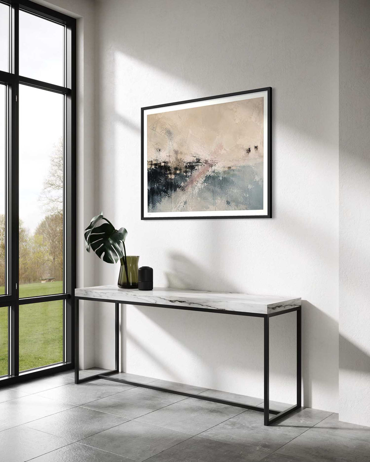 A framed abstract art print with beige, black, and blue tones hangs on a white wall above a modern console table in a bright, contemporary room with a large window.