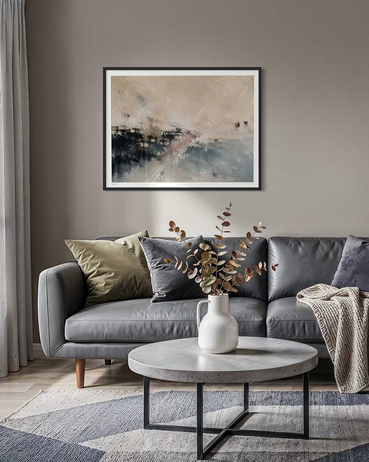 A framed abstract wall art print hangs above a gray sofa in a styled living room. The artwork features soft beige, gray, black, and muted pink tones.