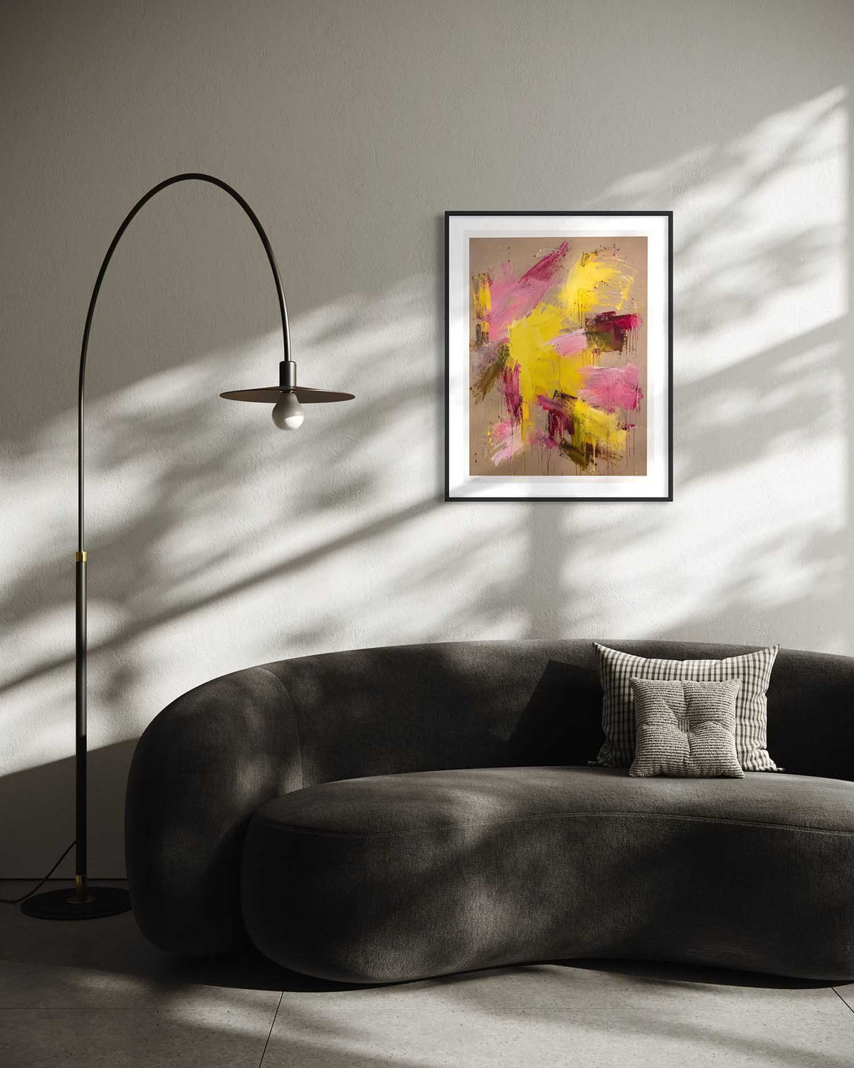 A framed abstract art print with yellow, pink, and brown brushstrokes hangs on a gray wall above a dark gray curved sofa in a modern living room setting with a black floor lamp and dramatic window shadows.
