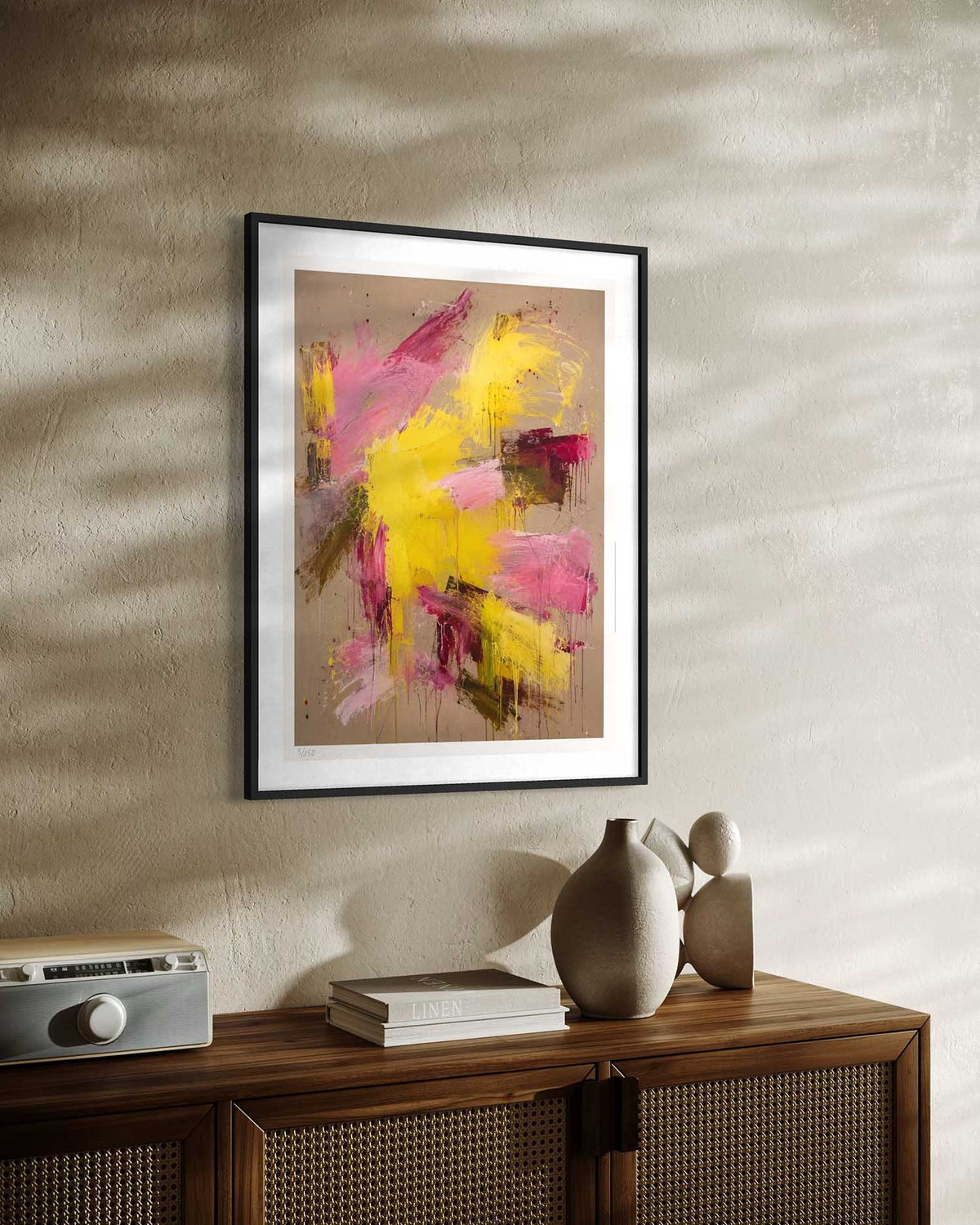 A framed abstract art print featuring bold strokes of yellow, pink, and brown on a beige background, styled on a textured beige wall above a wooden cabinet with decorative ceramic vases.