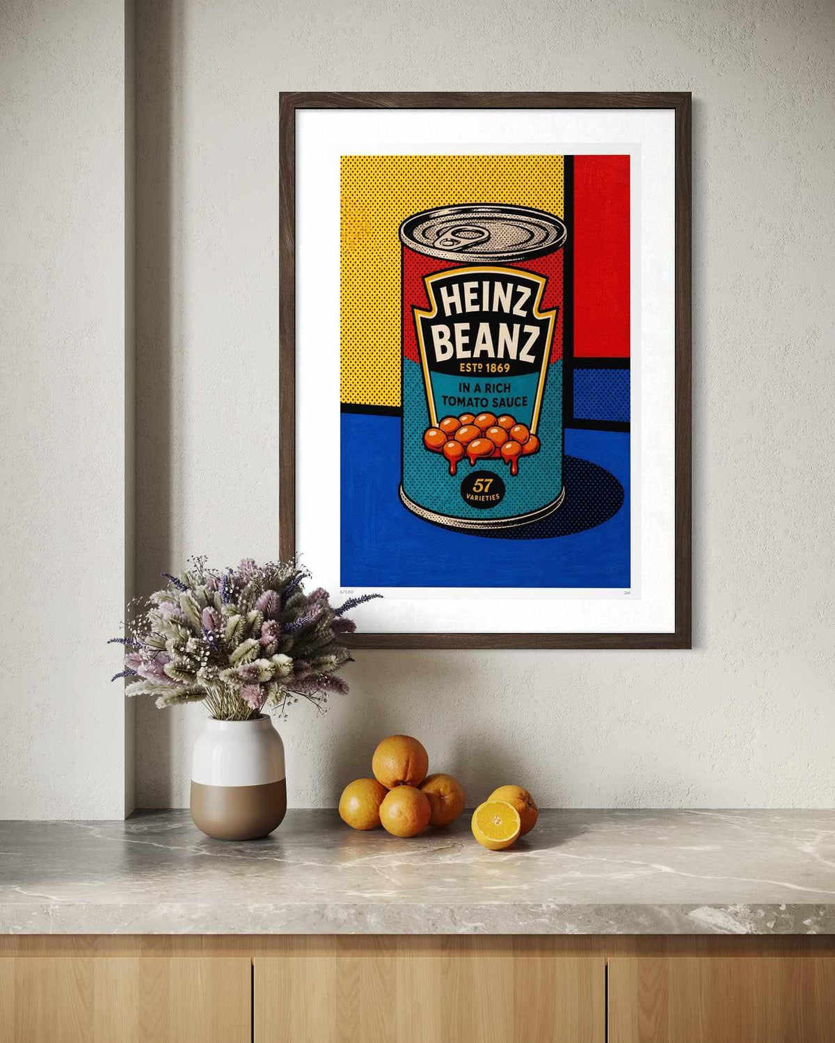 A framed pop-art print of a Heinz Bean product hanging on a light beige wall above a marble countertop with a vase of flowers and oranges.