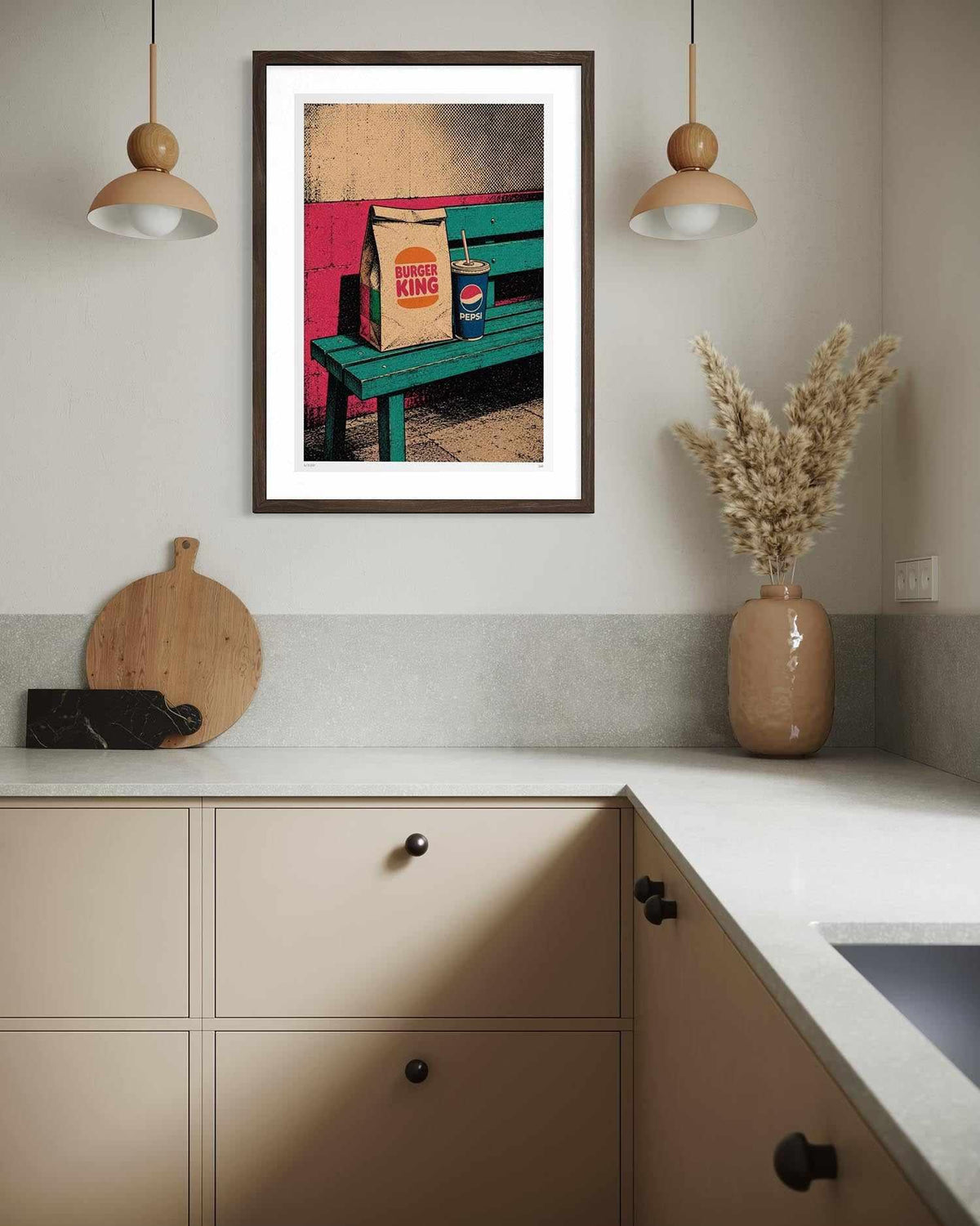 A framed art print featuring a colorful pop-art scene of a green bench with a Burger King bag and a Pepsi can, displayed on a kitchen wall above a beige countertop with decorative items.