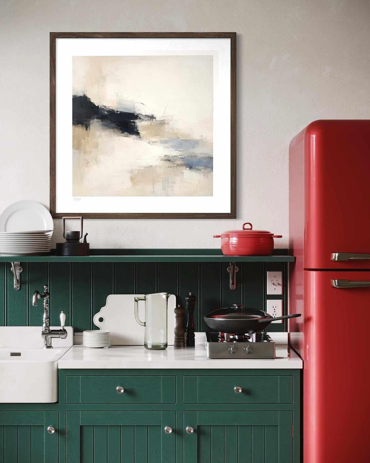 A framed abstract art print with beige, black, and blue tones hangs on a light gray wall above a green kitchen counter with a white sink, red refrigerator, and red pot.
