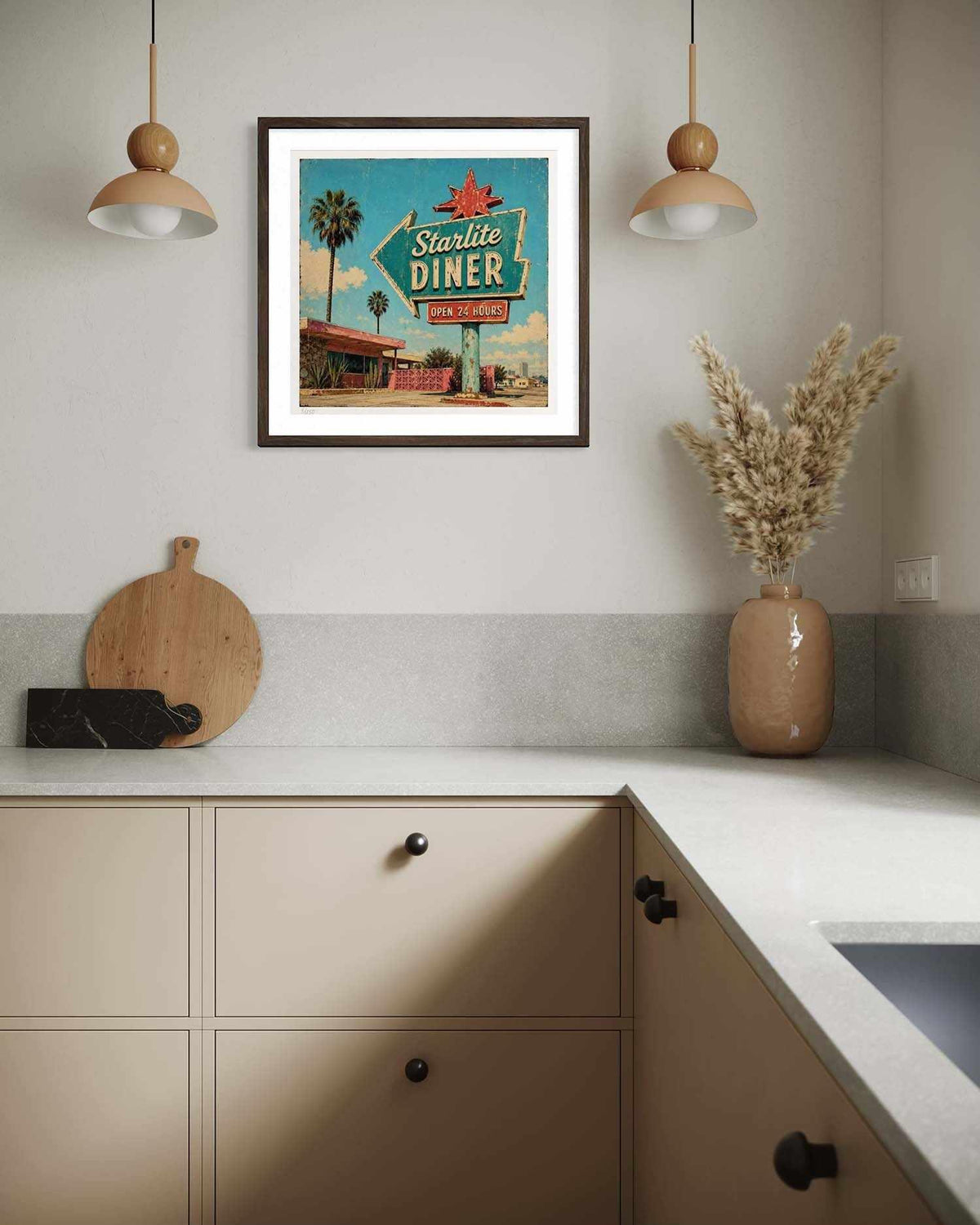 A framed art print featuring a retro 'Sterlite Diner' sign hangs on a kitchen wall above beige cabinets. The print has a vintage mid-century design with a teal sky, palm trees, and a diner, surrounded by a white mat and dark frame.
