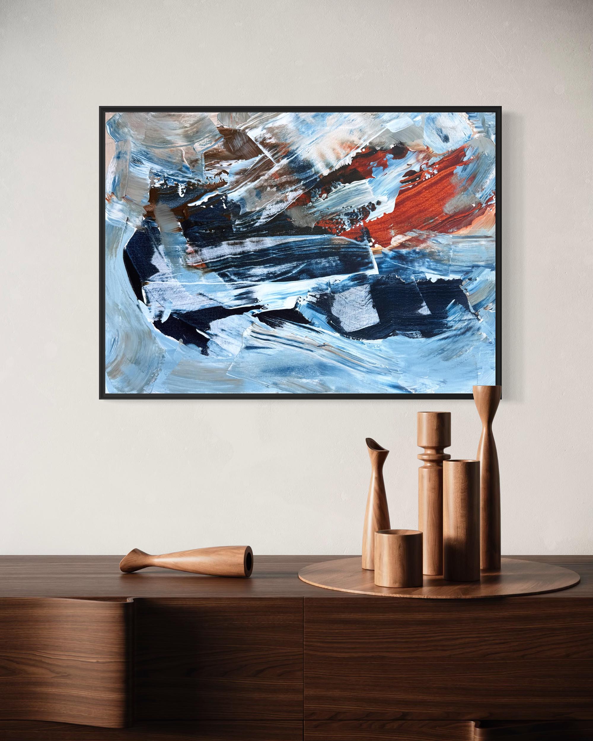 An abstract framed painting with blue, white, black, and red brushstrokes, displayed on a light beige wall above a dark wood console table with decorative wooden objects.