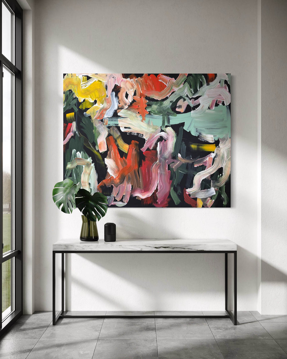 An abstract canvas painting with bold brushstrokes in red, pink, green, yellow, black, and white, displayed on a white wall above a marble-top console table with a green plant in a vase.