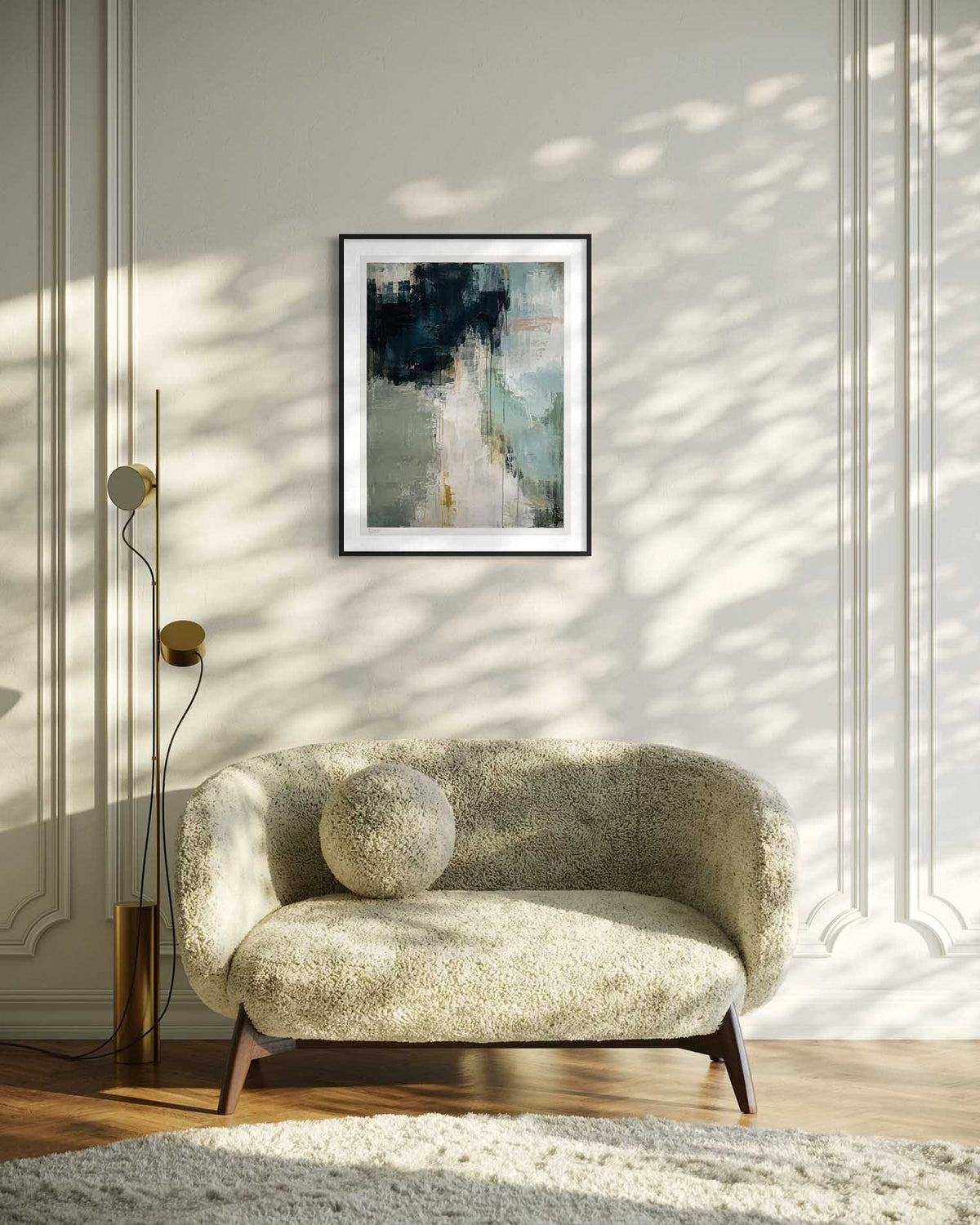 A framed abstract art print with navy, gray, and beige tones hangs on a paneled wall above a textured beige armchair in a sunlit room with leaf shadows.