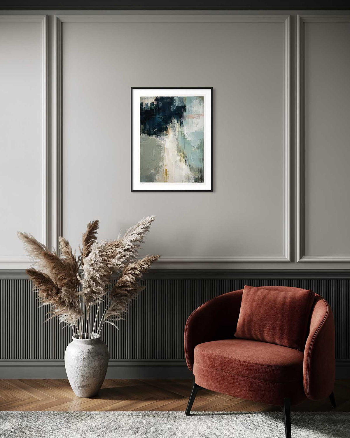 A framed abstract art print hangs on a gray paneled wall above a rust-red armchair and a vase of dried pampas grass in a styled interior room.
