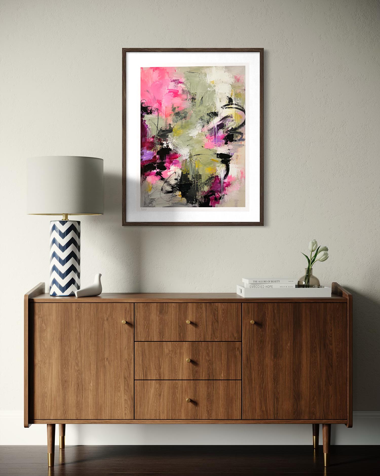 A framed abstract art print featuring vibrant pink, magenta, black, and olive green brushstrokes on a light gray background, styled in a mid-century modern room above a wooden sideboard with a lamp and books.
