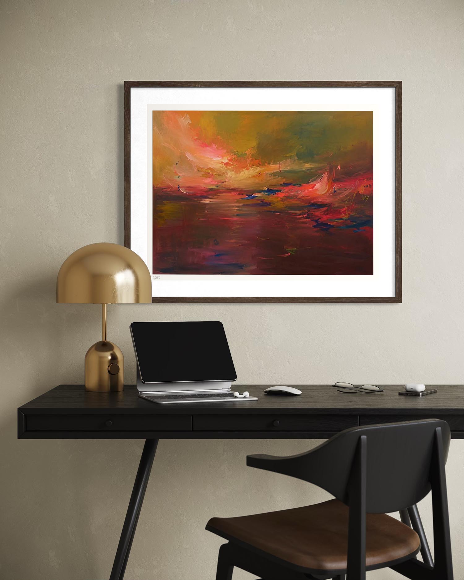 A framed abstract landscape painting with vibrant red, orange, yellow, and green brushstrokes, displayed on a beige wall above a black desk with a laptop, gold lamp, and chair.