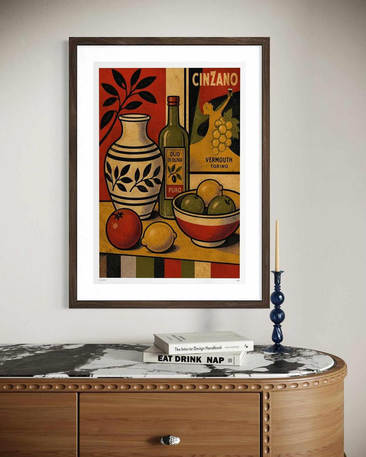 A framed vintage-style still life art print hangs on a light gray wall above a wooden console table with a marble top. The artwork depicts a vase, bottle, fruit, and a sign reading CINZANO.