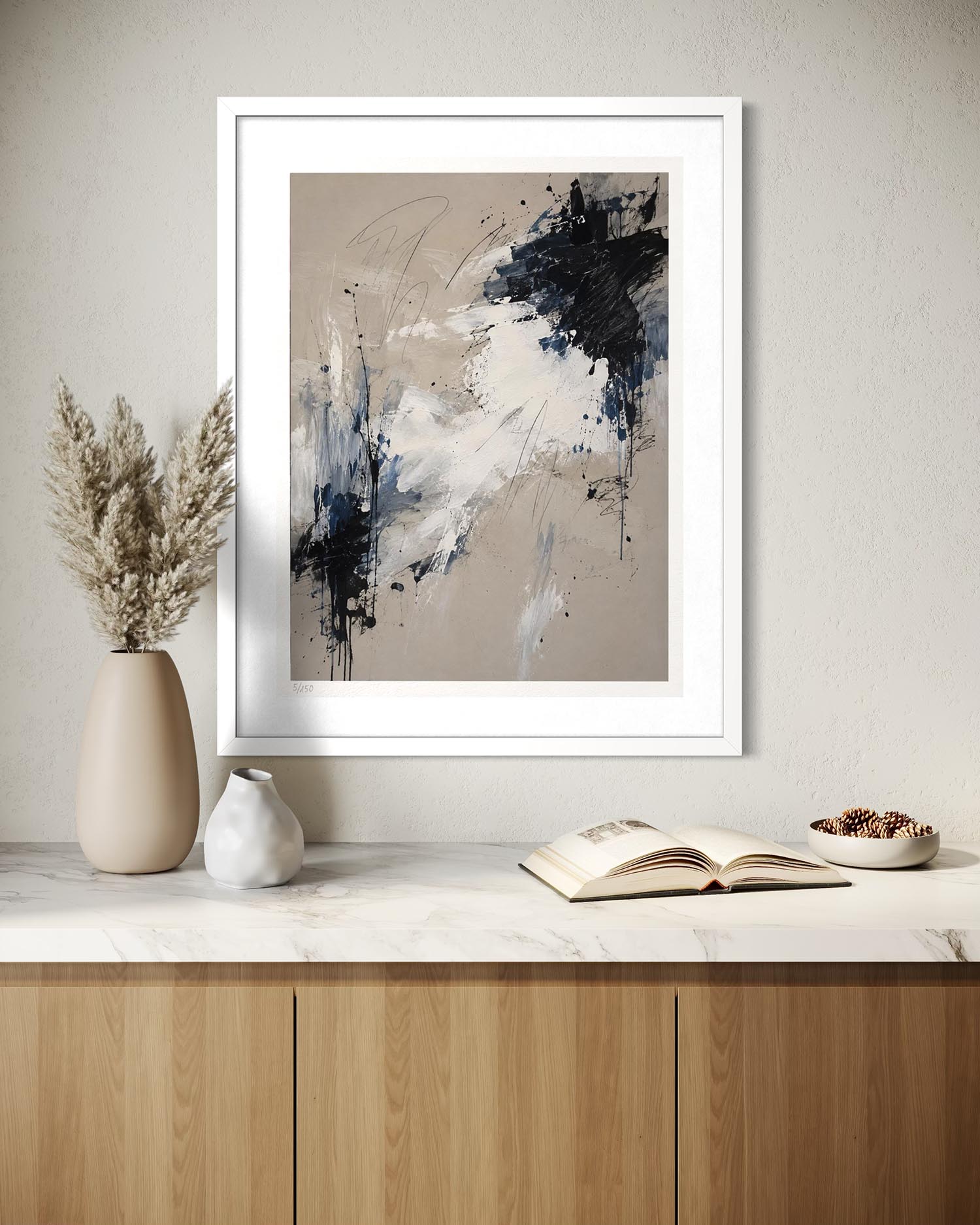 A framed abstract art print with a white frame hangs on a textured beige wall above a marble countertop with a wooden cabinet. The print features beige, white, and dark navy/black brushstrokes.