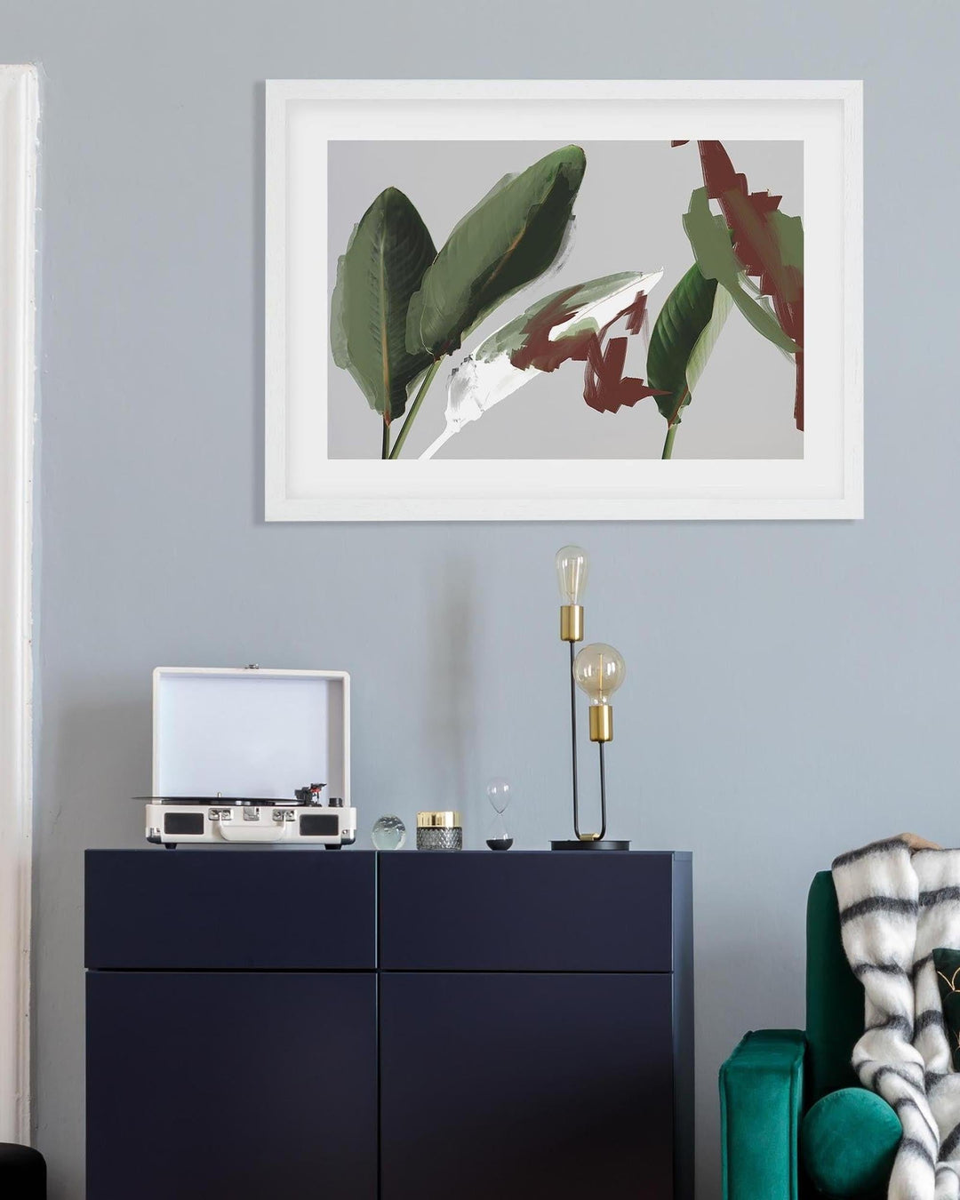 Framed artwork of leaves on a wall above a dark blue cabinet with decorative items.