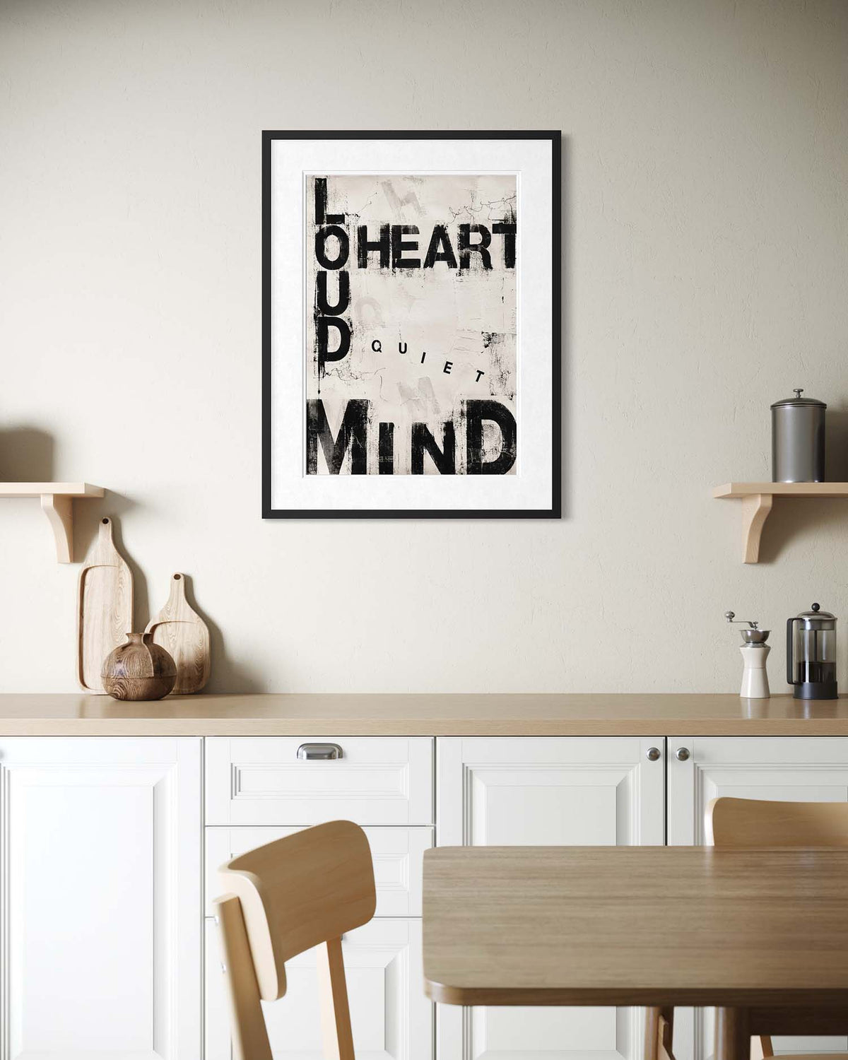 A framed typographic wall art print hangs on a beige wall above a kitchen counter. The print features black text reading 'LOUD HEART QUIET MIND' over a light textured background, enclosed in a black frame with a white mat.
