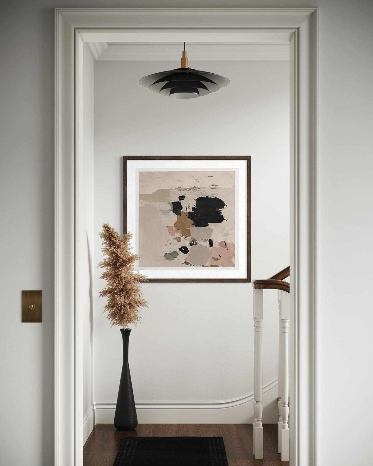 A framed abstract art print with beige, black, and brown tones hangs on a white wall in a hallway, with a black vase of dried grass and a black pendant light above.