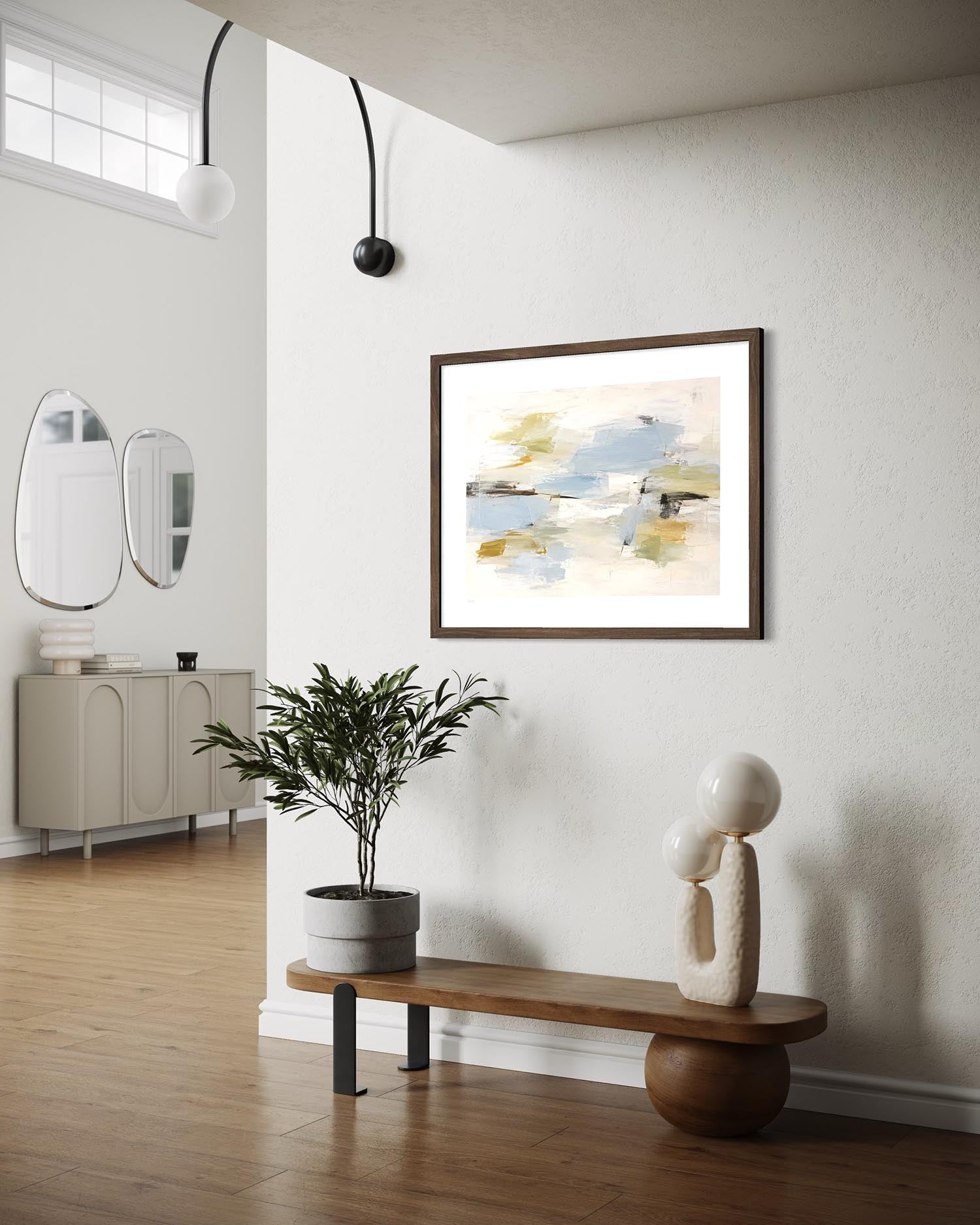 A framed abstract art print with soft blue, beige, and yellow brushstrokes hangs on a white wall in a modern, sunlit entryway with a wooden bench, potted plant, and sculptural decor.