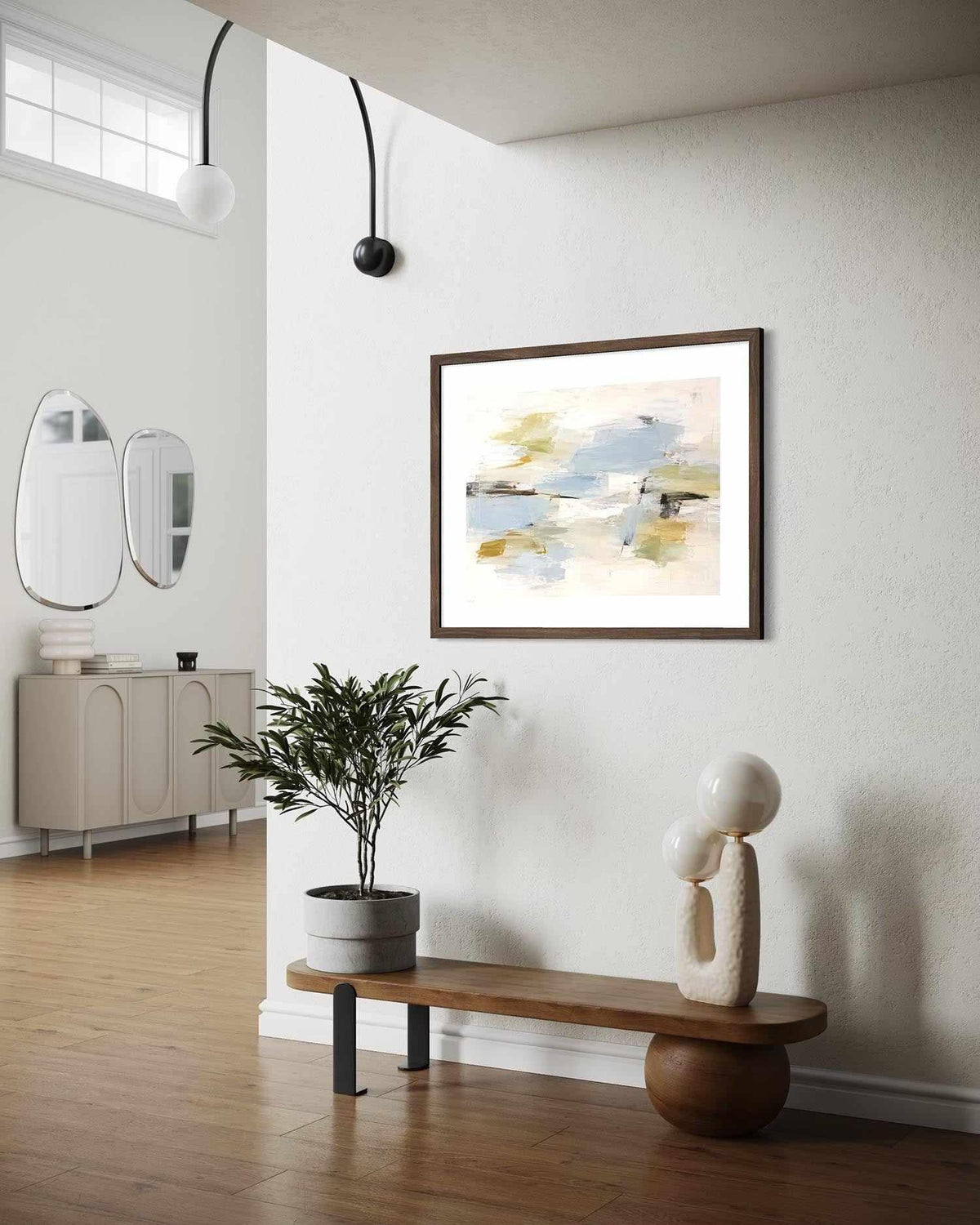 A framed abstract art print with soft blue, beige, and yellow brushstrokes hangs on a white wall in a modern, sunlit entryway with a wooden bench, potted plant, and sculptural decor.