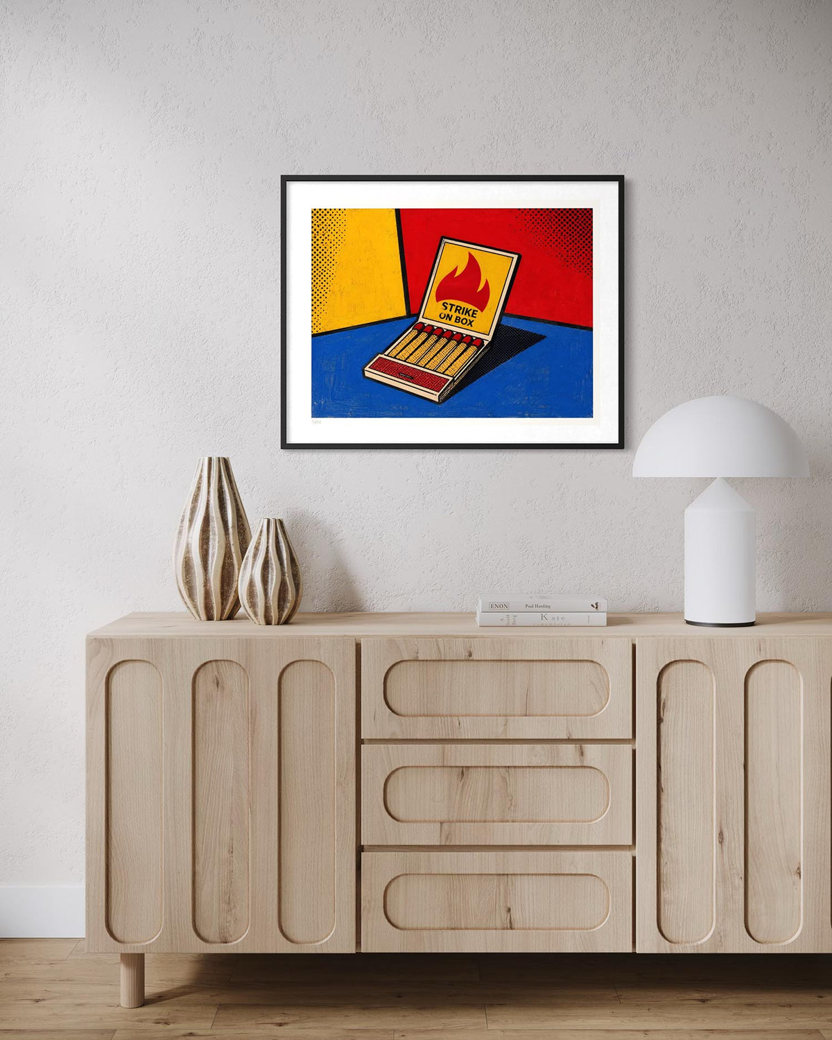 A framed pop-art print of a matchbox with bold red, yellow, and blue colors hangs on a light gray wall above a light wood sideboard with decorative vases and a white lamp.