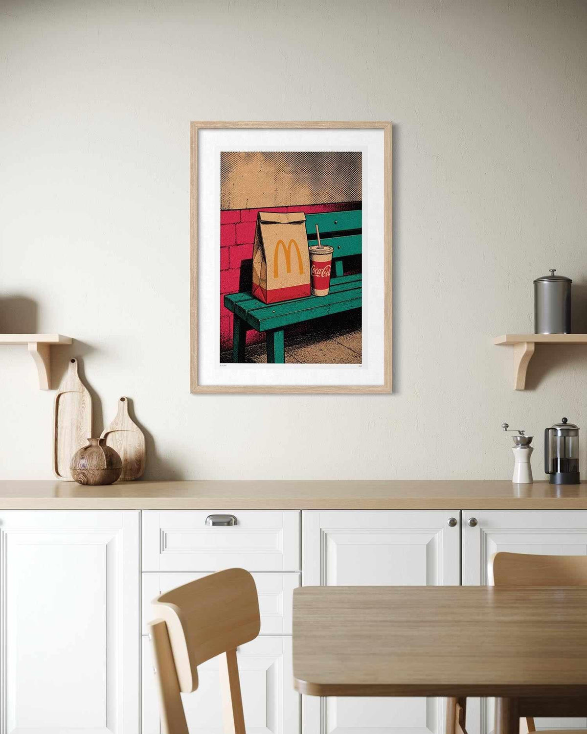A framed art print featuring a pop-art style illustration of a McDonald's bag and drink on a green bench against a red brick wall, displayed on a beige wall above a kitchen counter.