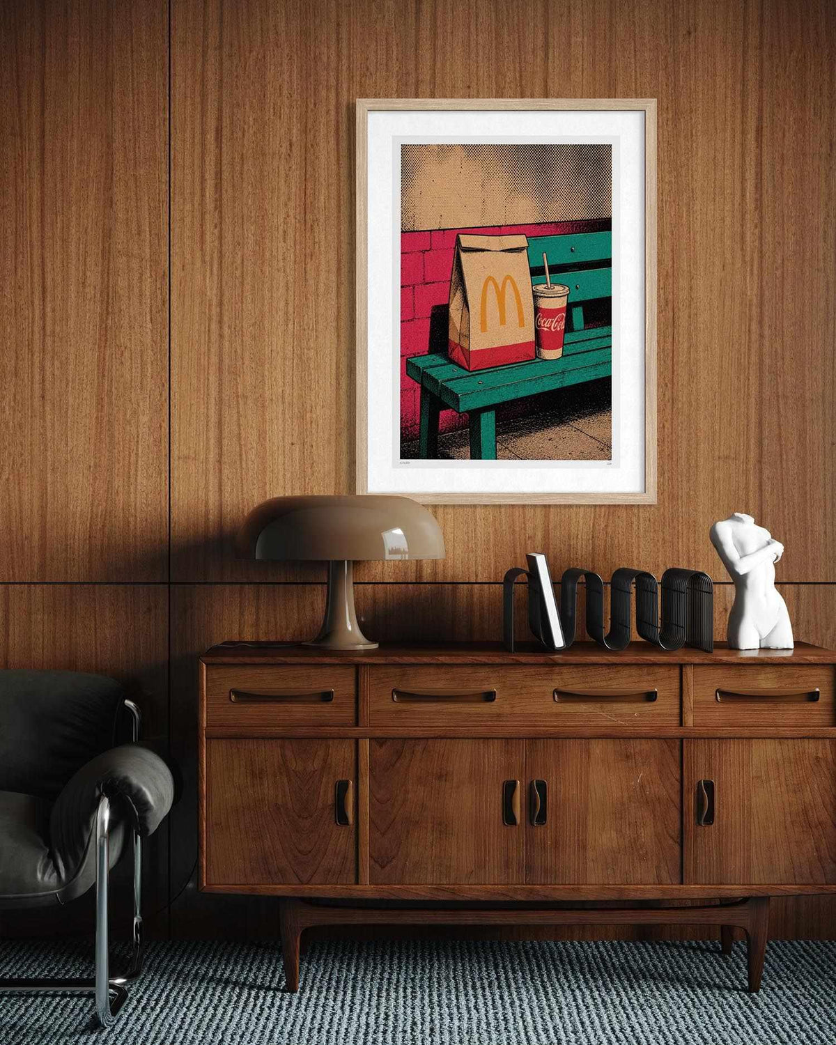 A framed art print featuring a colorful pop-art scene of a McDonald's bag and drink on a green bench, displayed on a wood-paneled wall above a wooden credenza in a modern room setting.