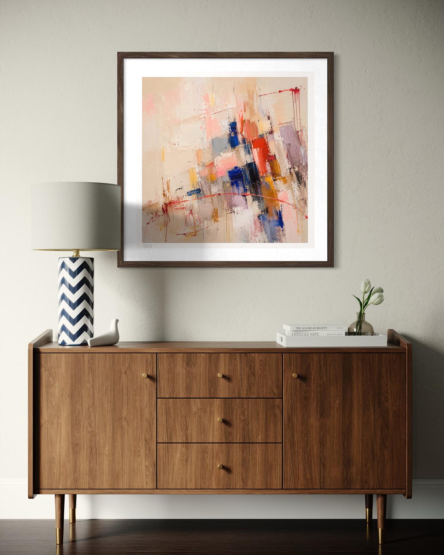 An abstract framed wall art print hangs above a mid-century modern wooden sideboard in a styled room setting. The artwork features a multicolor abstract design with brushstrokes of blue, red, orange, and pink on a light beige background, enclosed in a dark brown frame with a white mat.