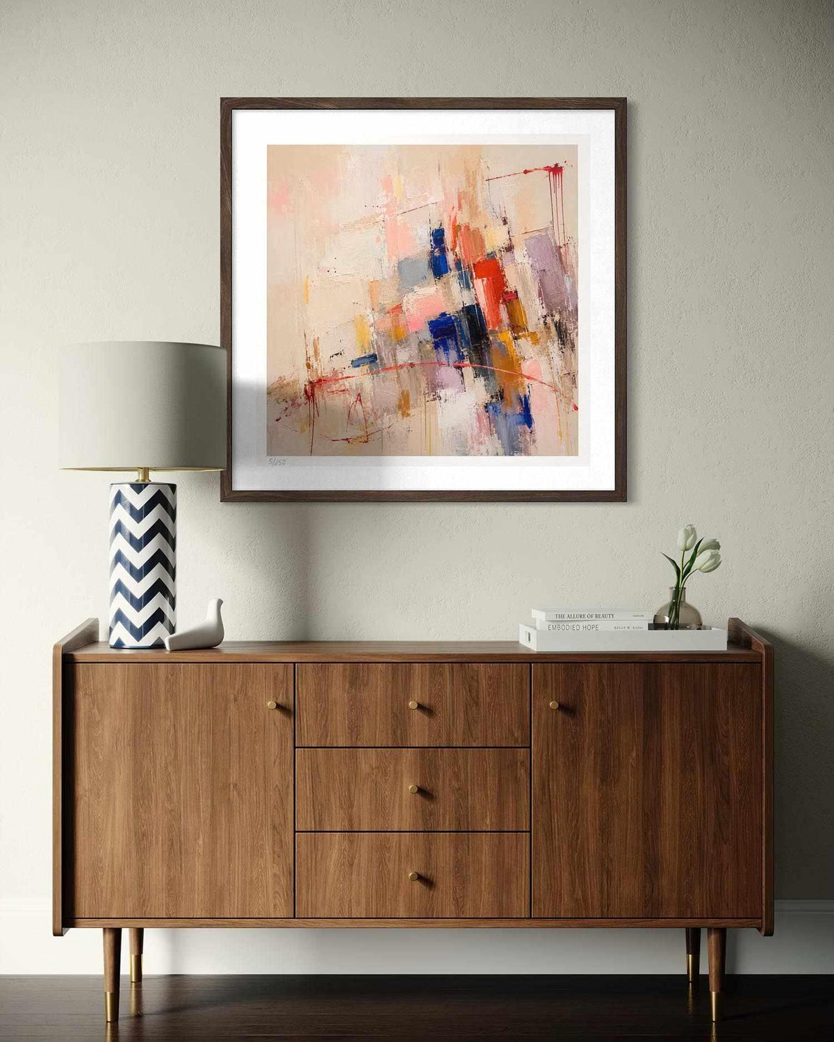 An abstract framed wall art print hangs above a mid-century modern wooden sideboard in a styled room setting. The artwork features a multicolor abstract design with brushstrokes of blue, red, orange, and pink on a light beige background, enclosed in a dark brown frame with a white mat.